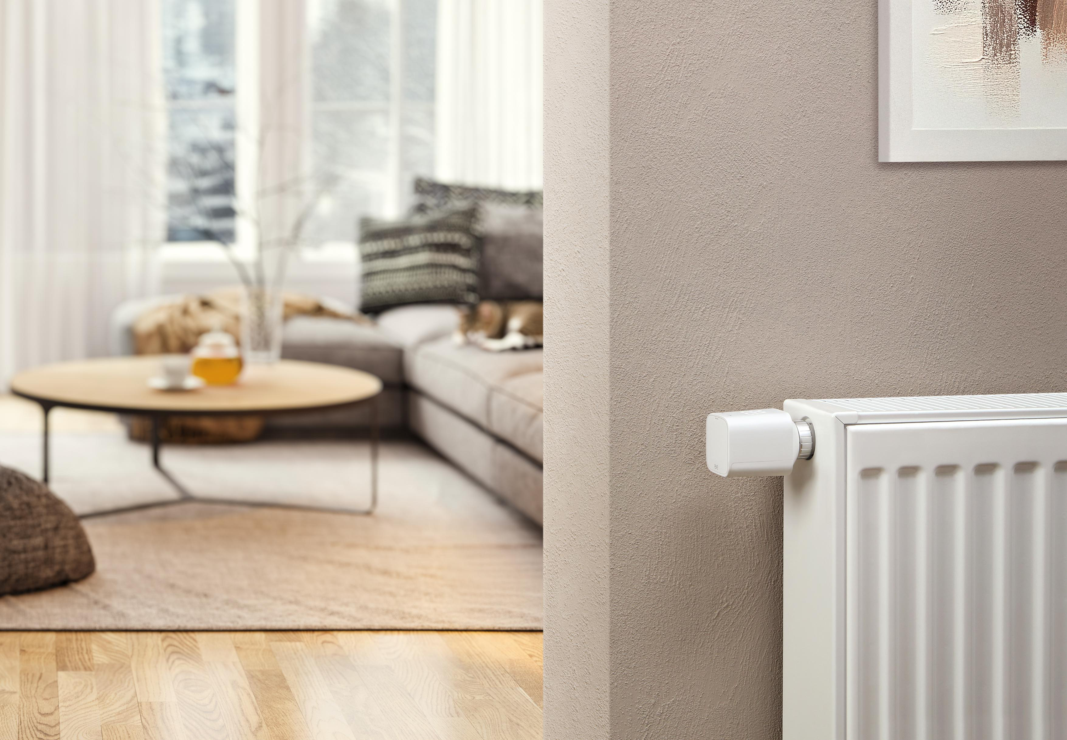 Wohnzimmerszene mit Heizkörper, Smart-Thermostat und einem Blick auf Möbel und Fenster.