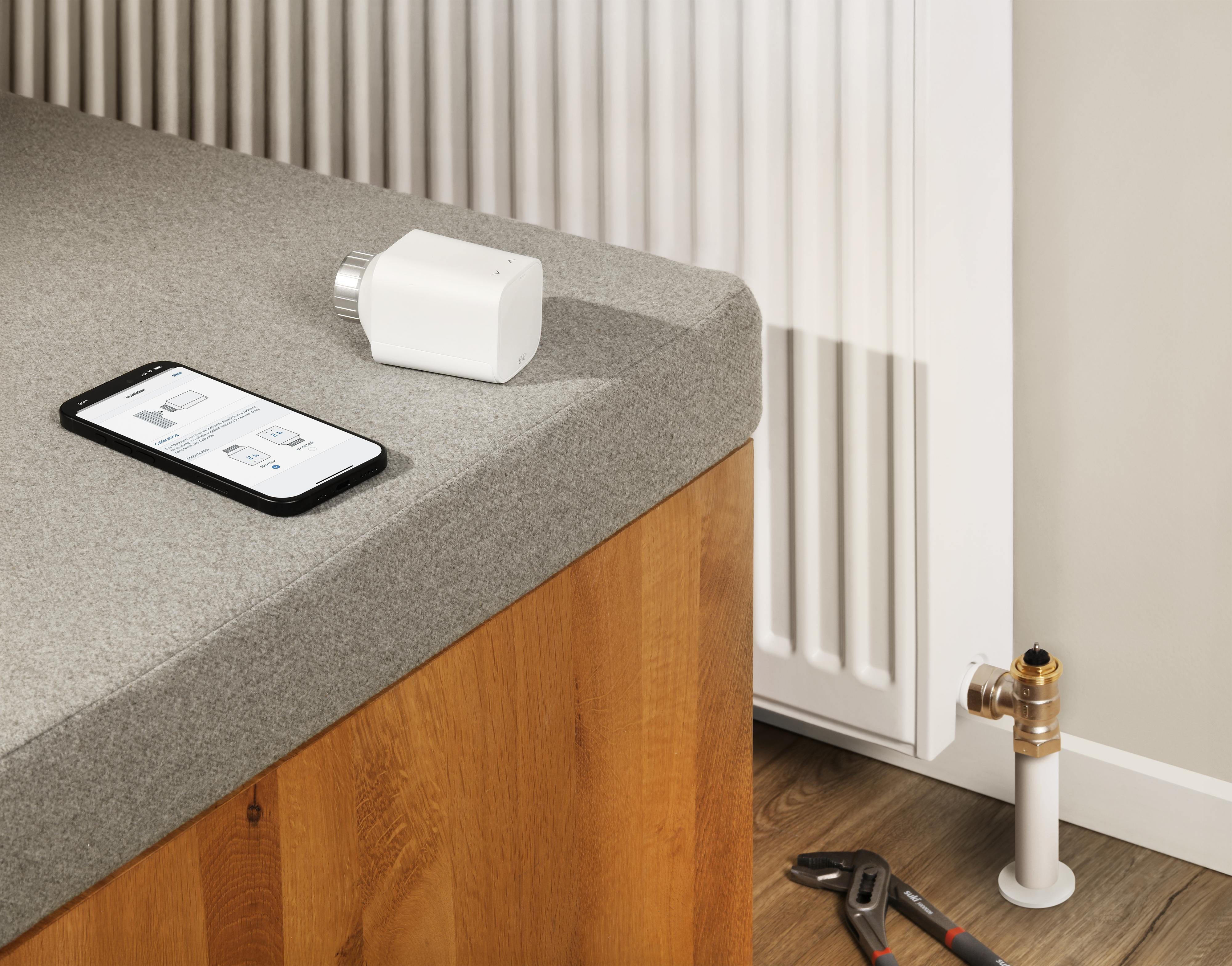 Ein weißer Thermostat und ein Telefon auf einer Bank. Das Telefon zeigt Installationsschritte. Heizkörper und Werkzeuge rechts.