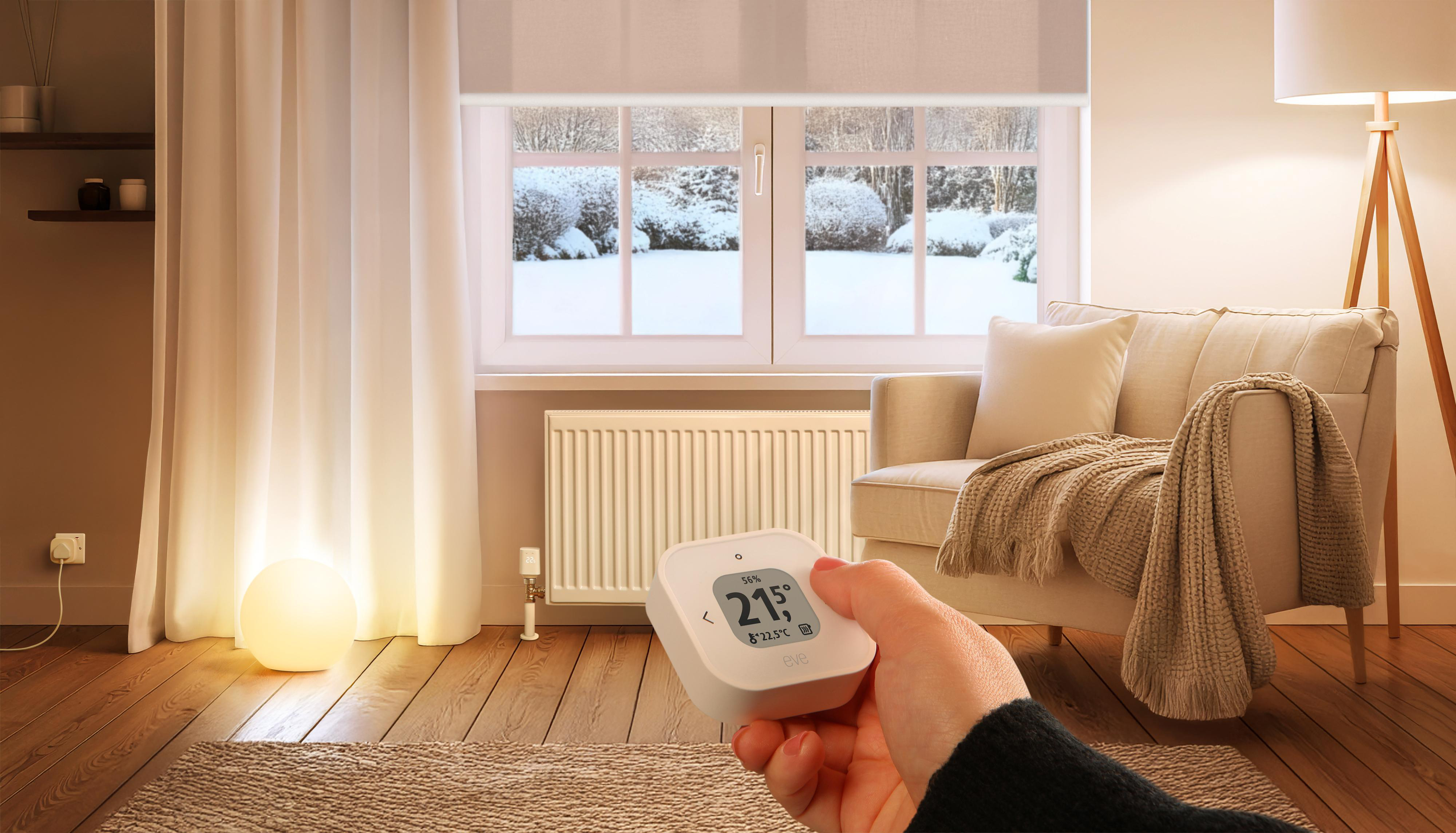 Eine Hand hält ein Thermostat in einem Raum. Das Display zeigt die Temperatur. Ein Fenster zeigt eine Schneelandschaft.