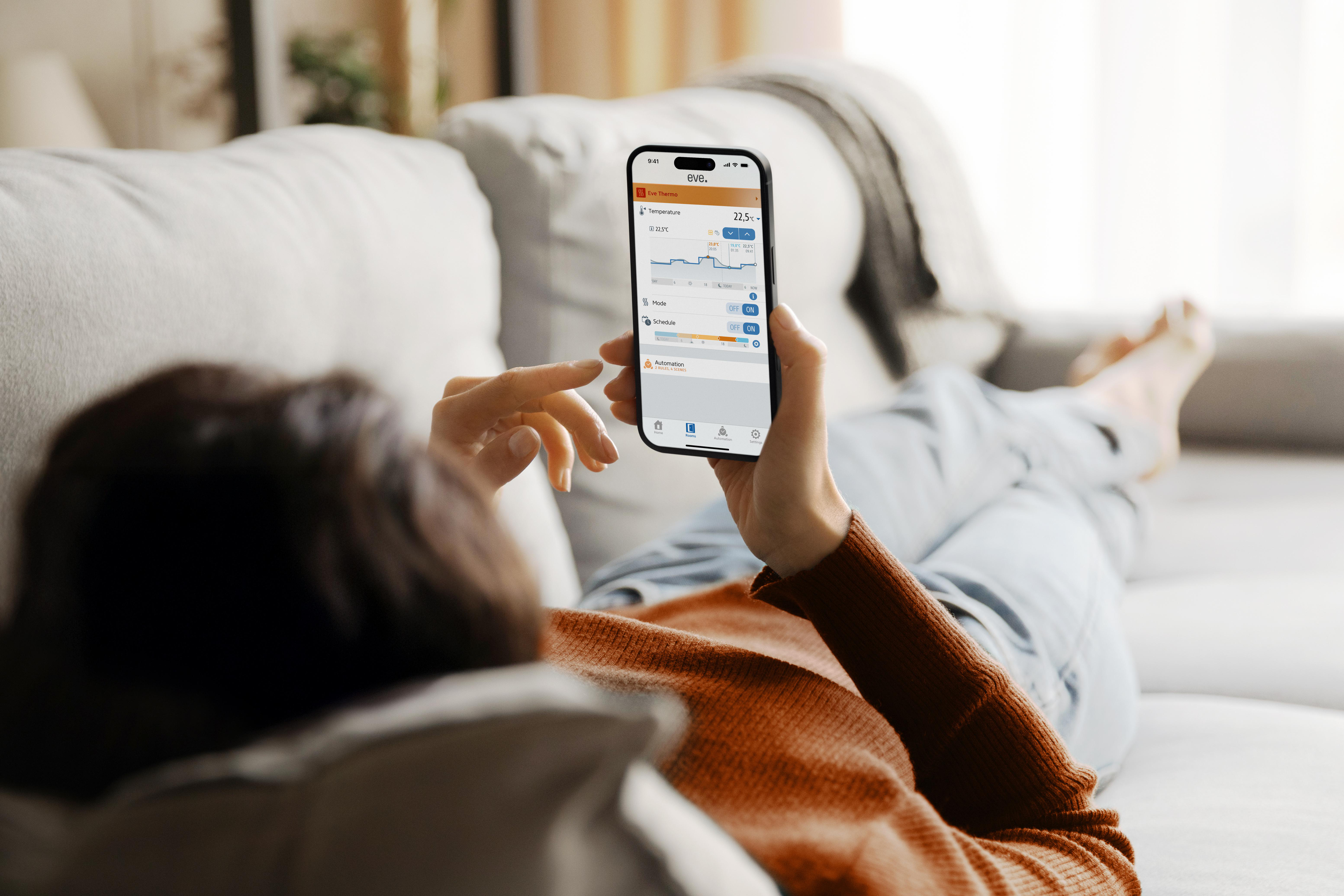 Eine Person liegt auf einem Sofa und hält ein Telefon, das eine Smart-Home-App-Oberfläche anzeigt.