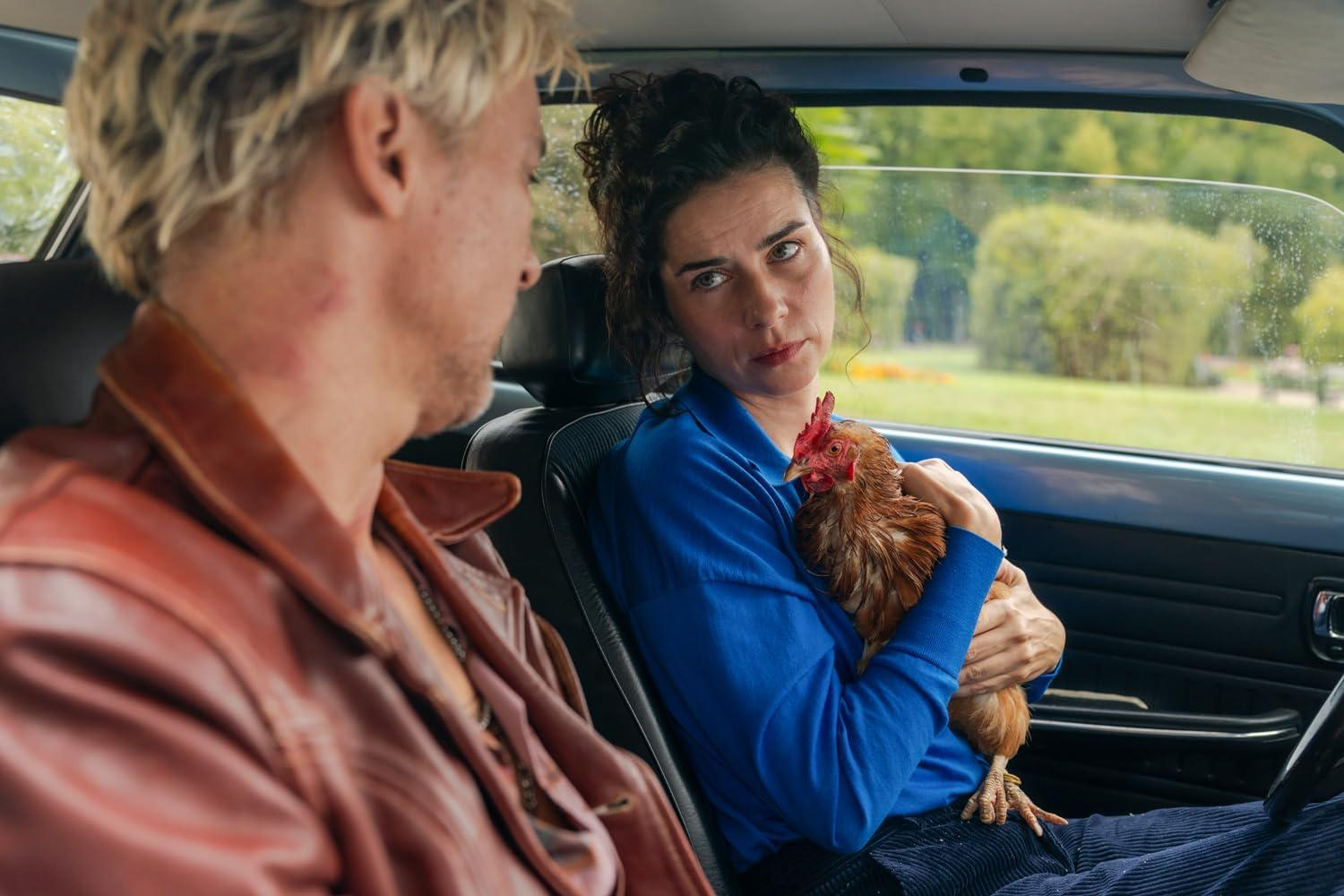 In einem Auto hält eine Frau ein Huhn. Ein Mann ist neben ihr. Im Hintergrund sind Bäume und ein blaues Auto zu sehen.