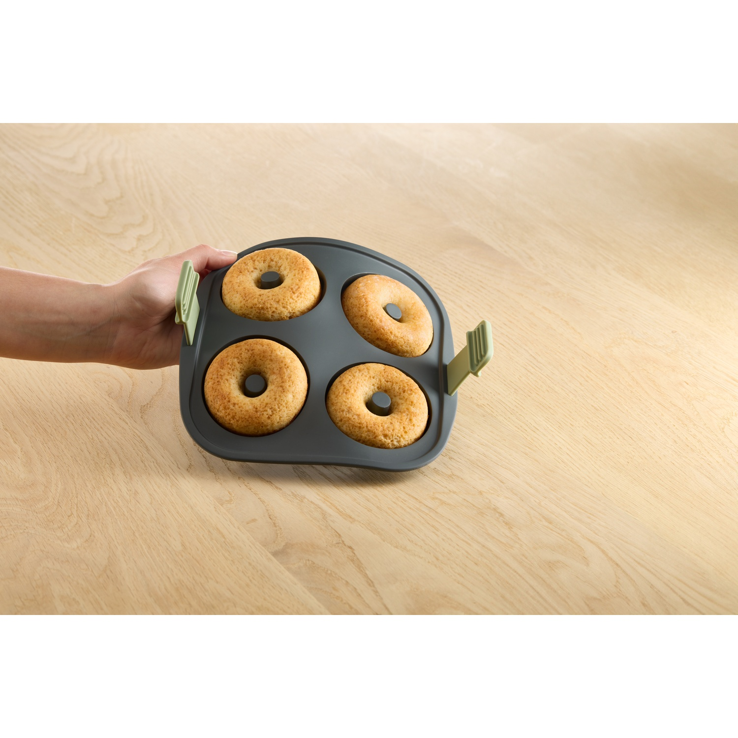 A hand holds a gray donut maker with four golden donuts. It rests on a wooden table.
