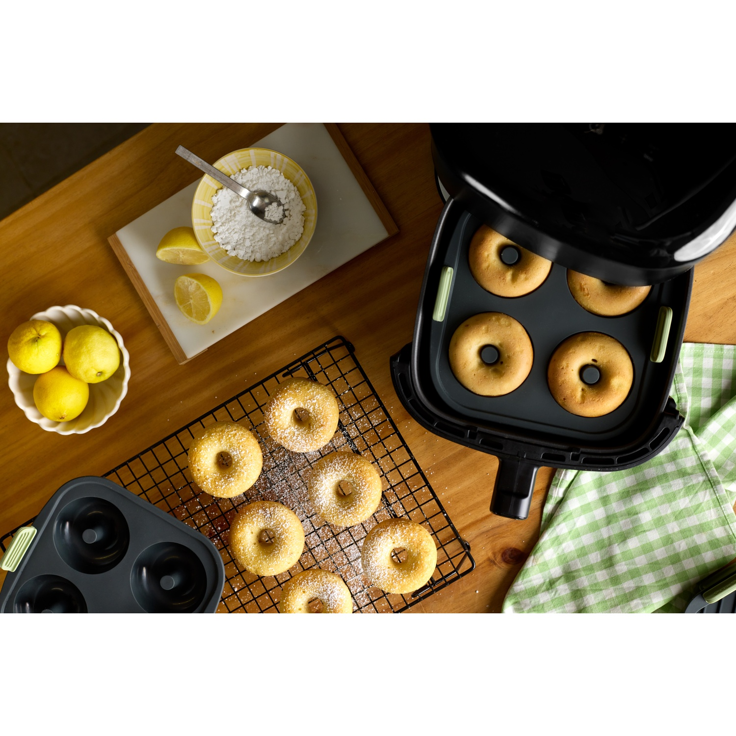 Overhead shot of donuts being made. Donuts on a rack, in an air fryer, and in a pan. Lemons, sugar, and a spoon are present.