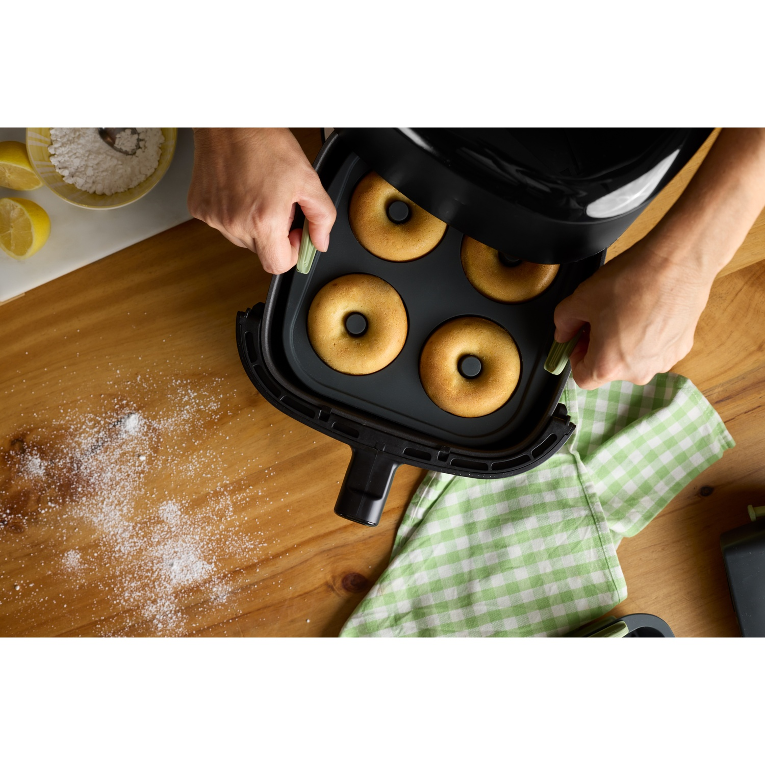 Hands holding an air fryer with four donuts. On a wooden table, lemons, flour, and a green checkered cloth are visible.