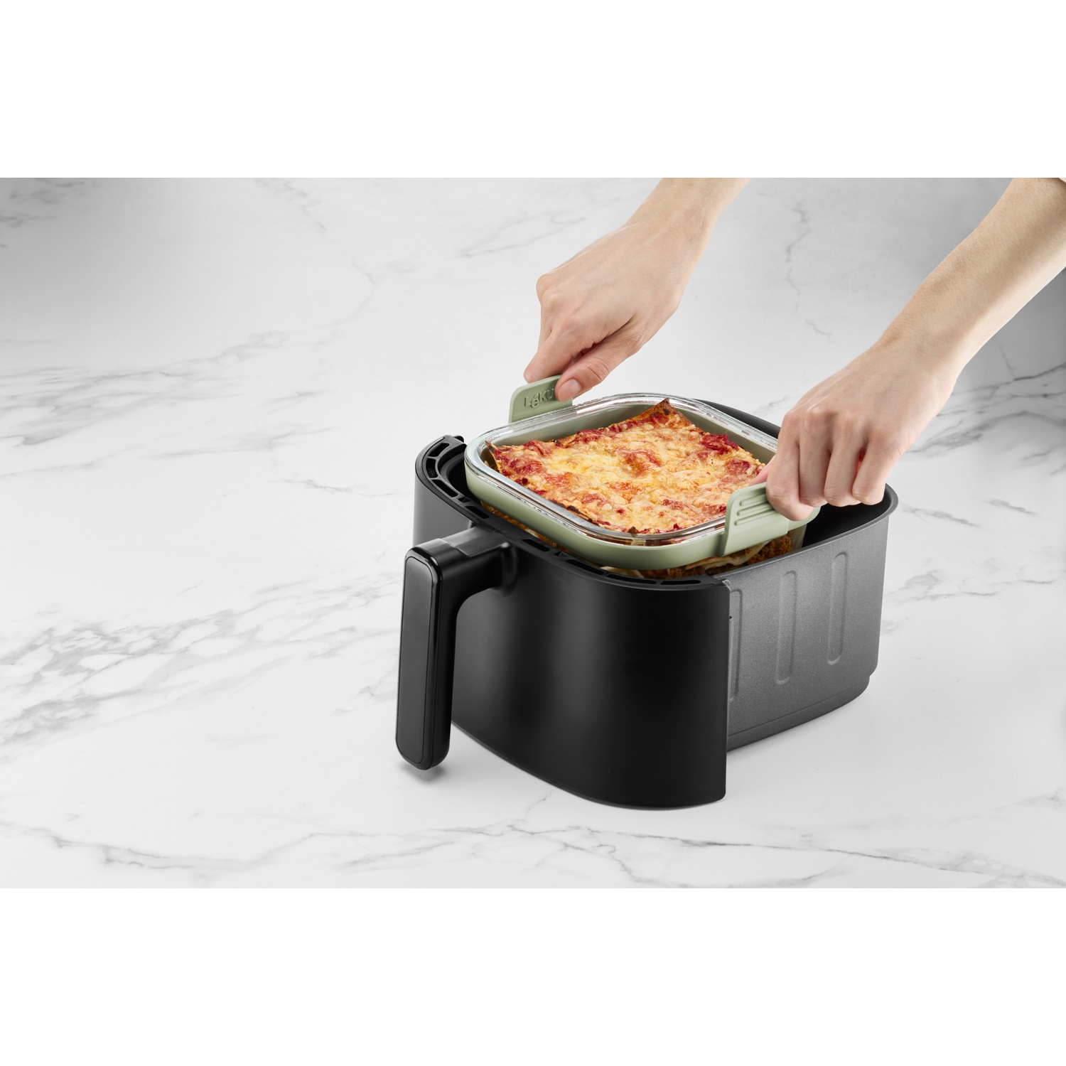 Hands placing a lasagna dish in a black air fryer. The lasagna is in a green dish, on a white marble surface.