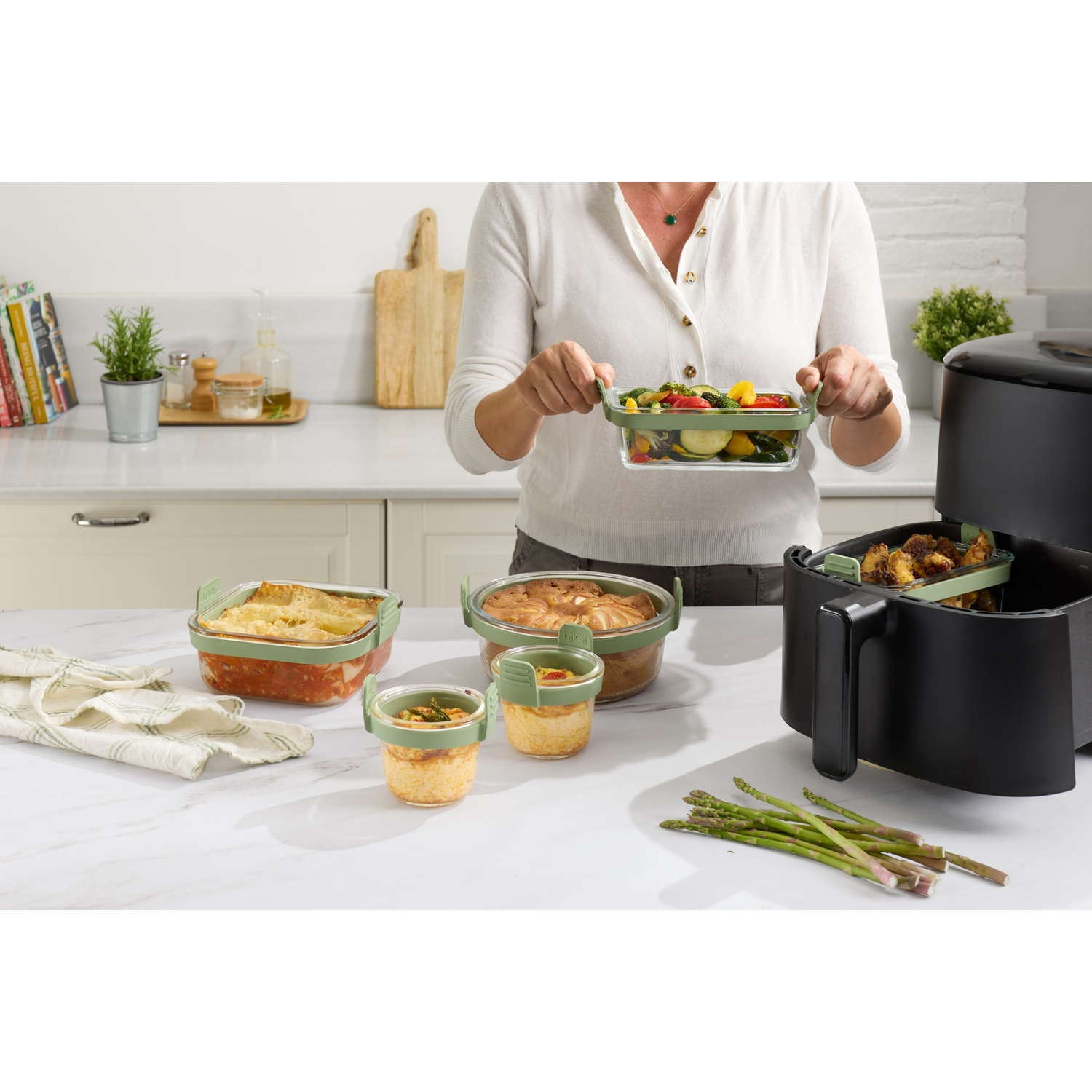 A woman holds a container of vegetables. Several containers of food are on the counter. An air fryer is on the right.