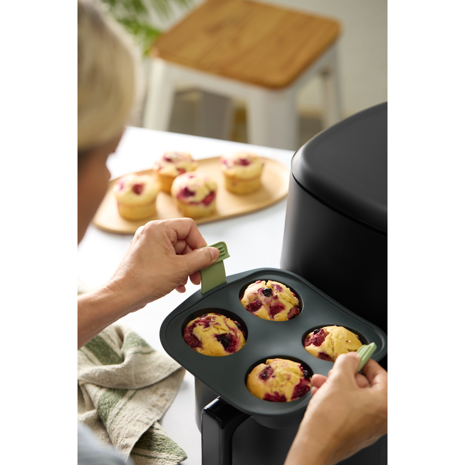 Une personne retire des muffins aux baies d'un plateau. D'autres muffins sont sur une assiette en bois. Une serviette verte est à proximité.