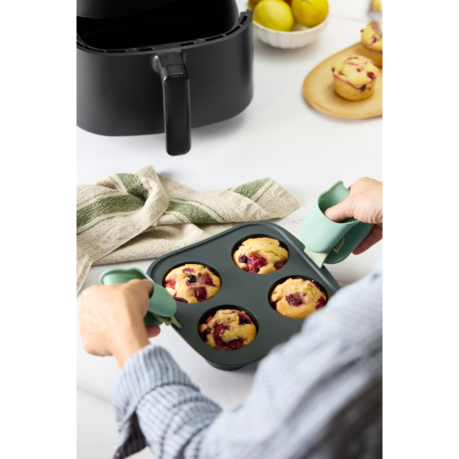 Mains tenant un plateau de muffins. En arrière-plan, un airfryer et un bol de citrons. Une planche en bois avec des muffins est également présente.