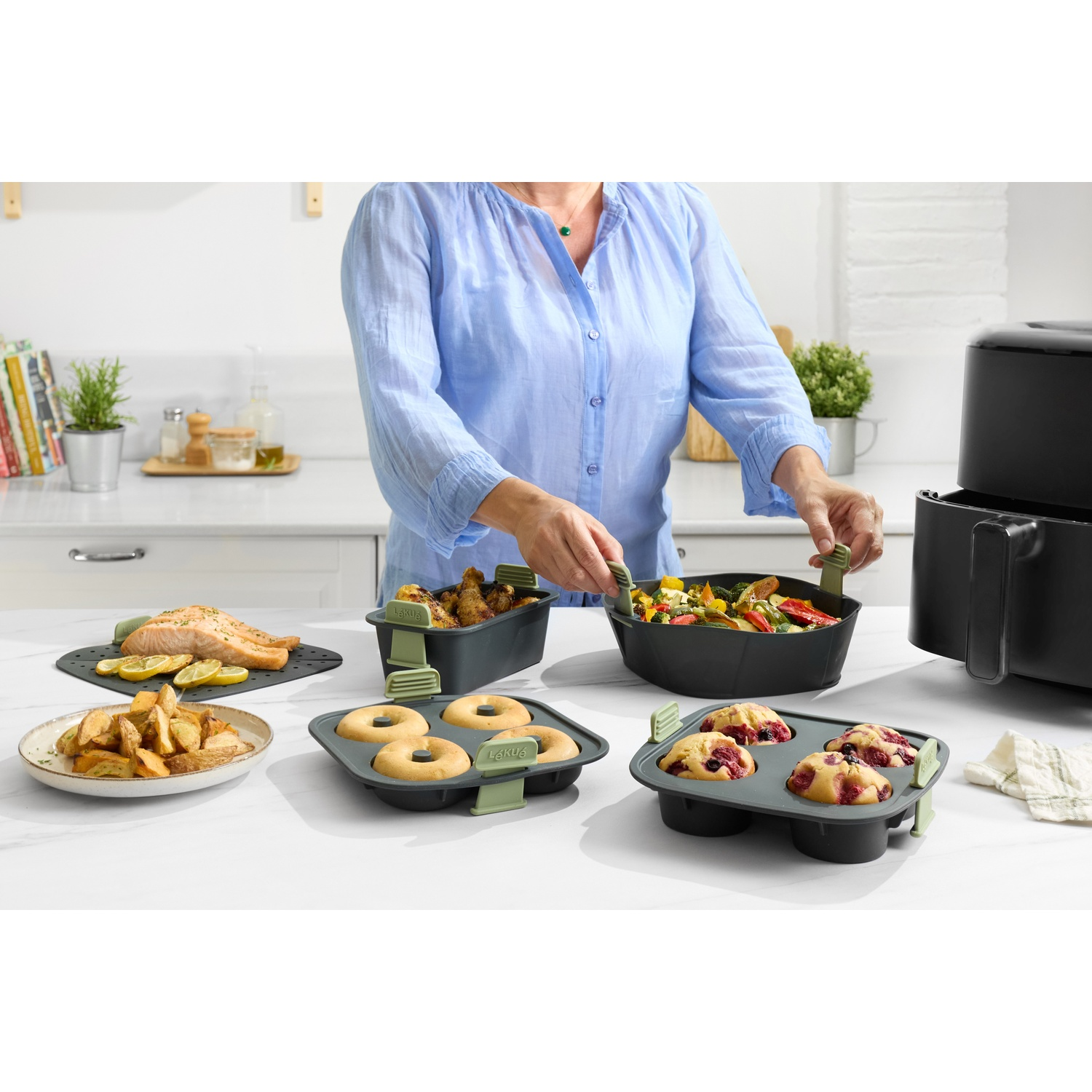Une personne prépare des aliments avec une friteuse à air. Des plats avec du saumon, des frites, des beignets, des muffins et des légumes sont sur la table.