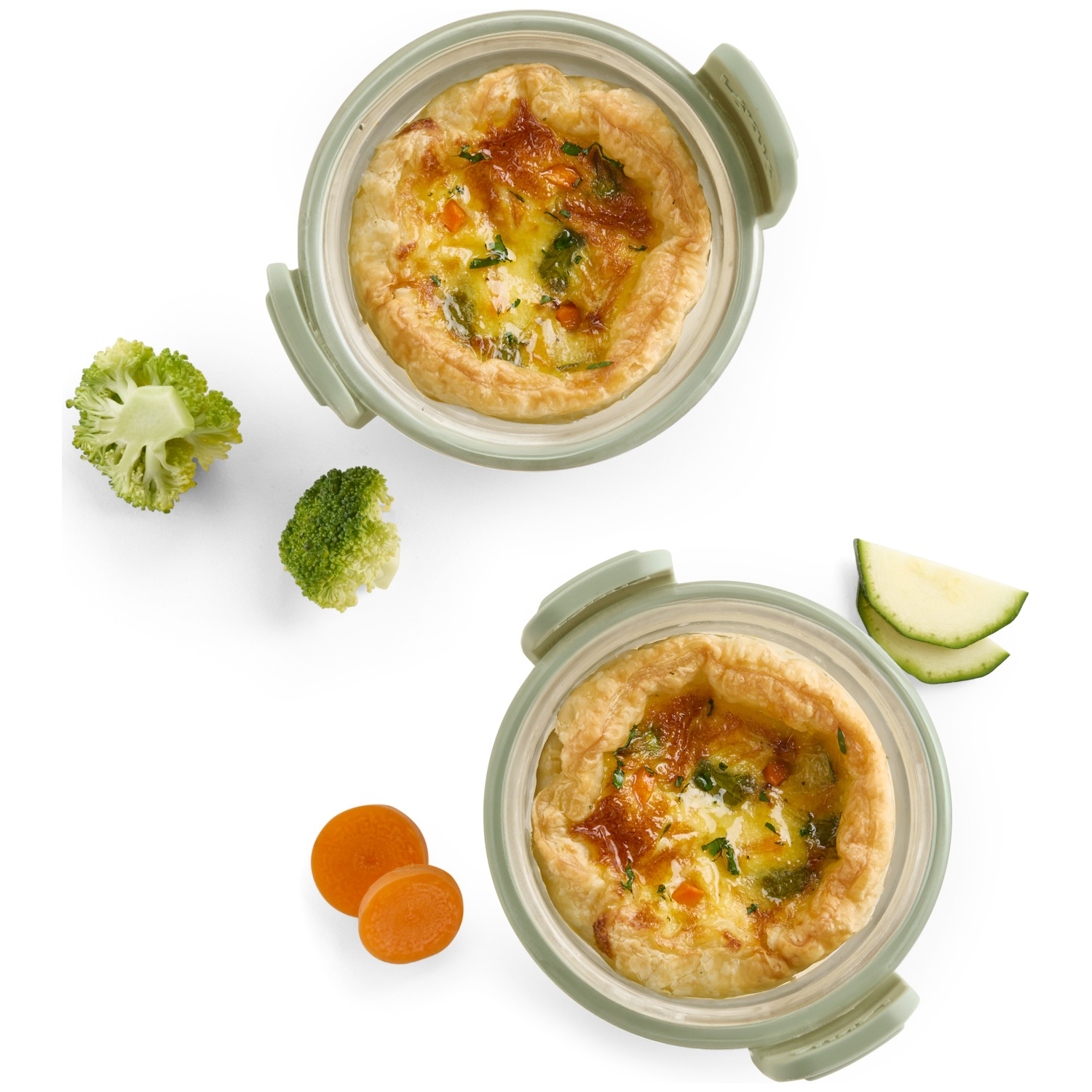 Twee quiches in groene kommen met groenten. Broccoli, wortelen en plakjes courgette verspreid op een wit oppervlak.
