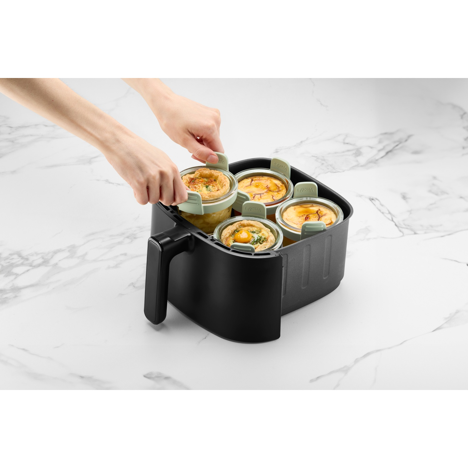 Handen plaatsen glazen potten met voedsel in een zwarte airfryer. Potten hebben groene deksels. Achtergrond is marmer.