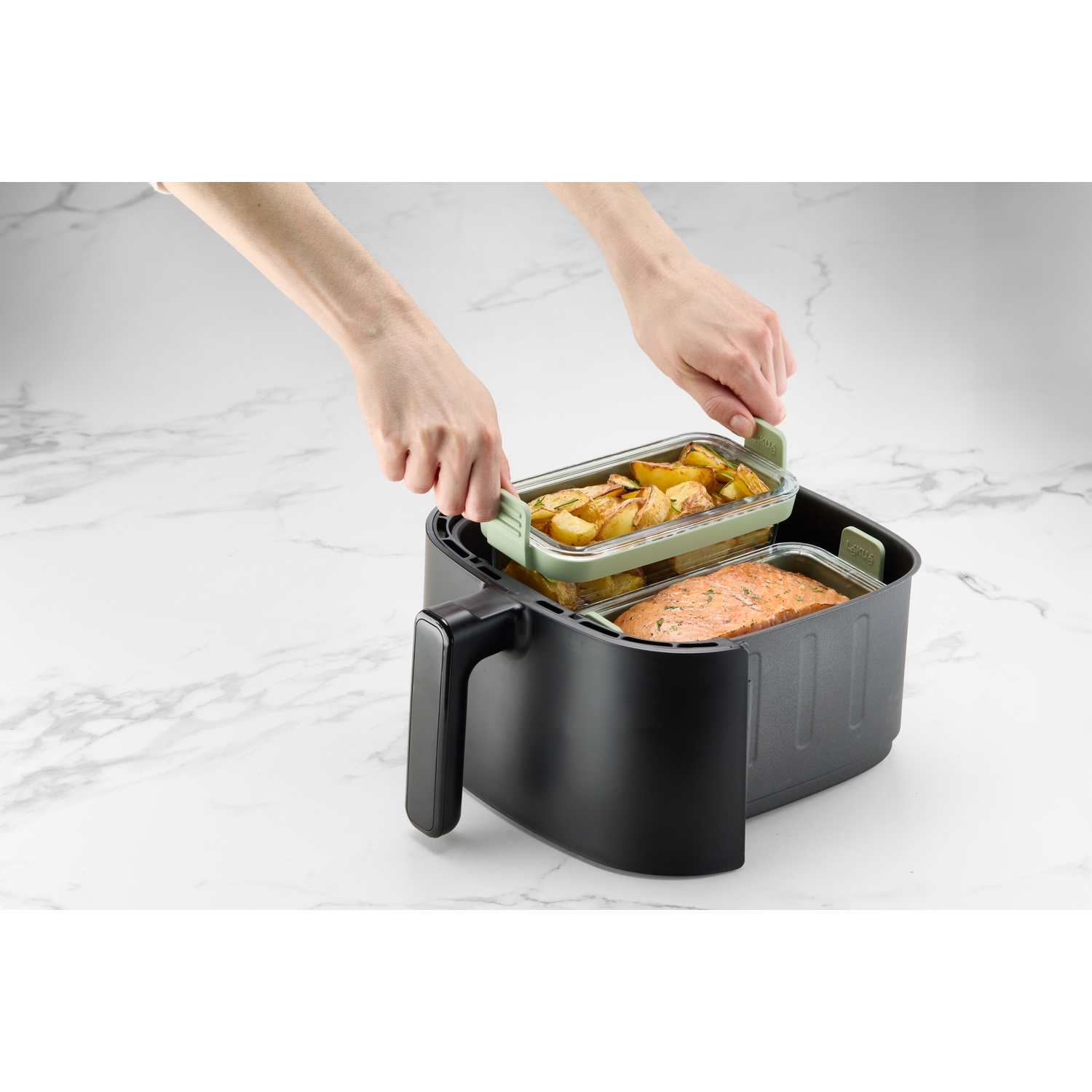 Hands placing a glass container with potatoes into a black air fryer. Another container with salmon is below.