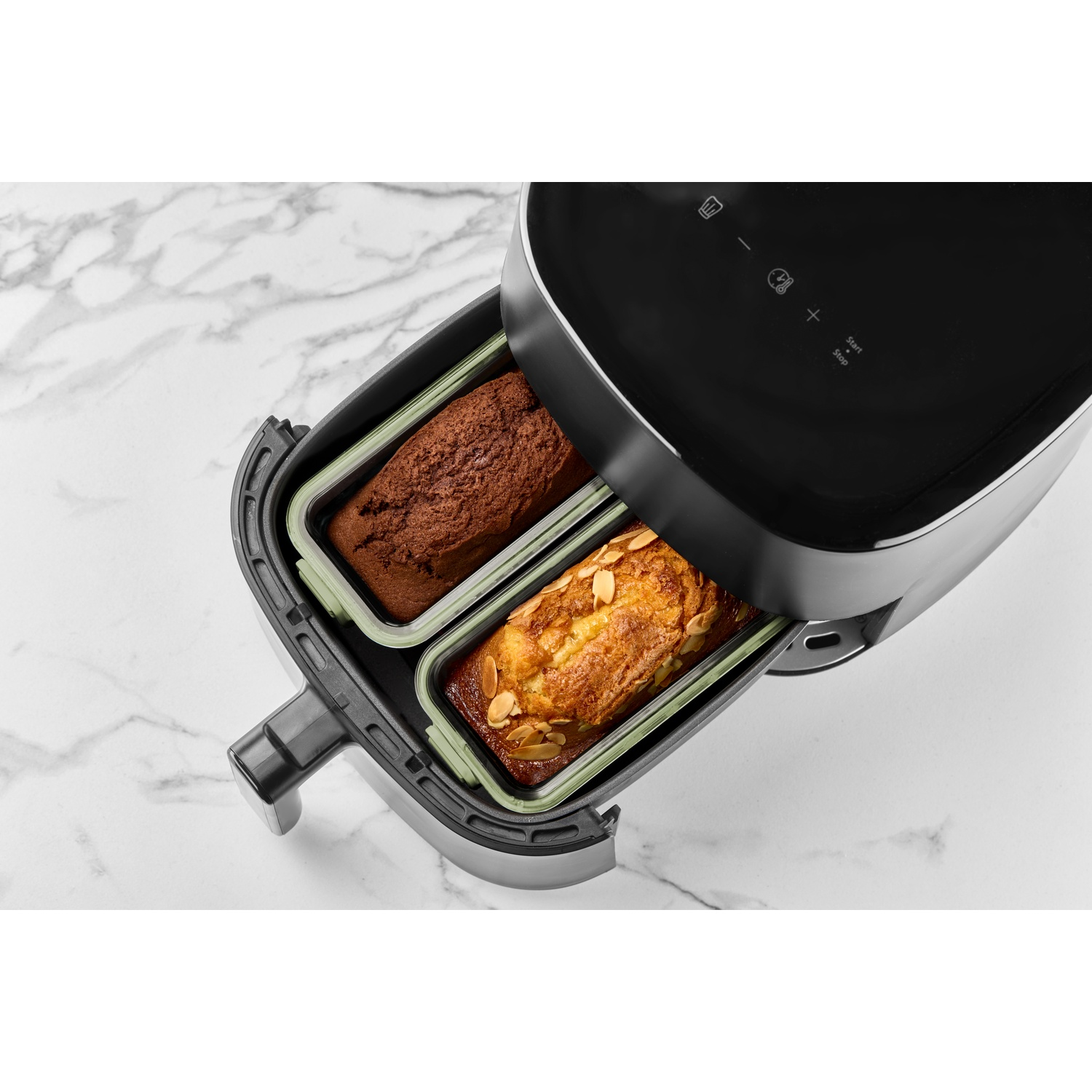 An air fryer with two loaf cakes inside. One is chocolate, the other is a golden cake with almonds. White marble background.