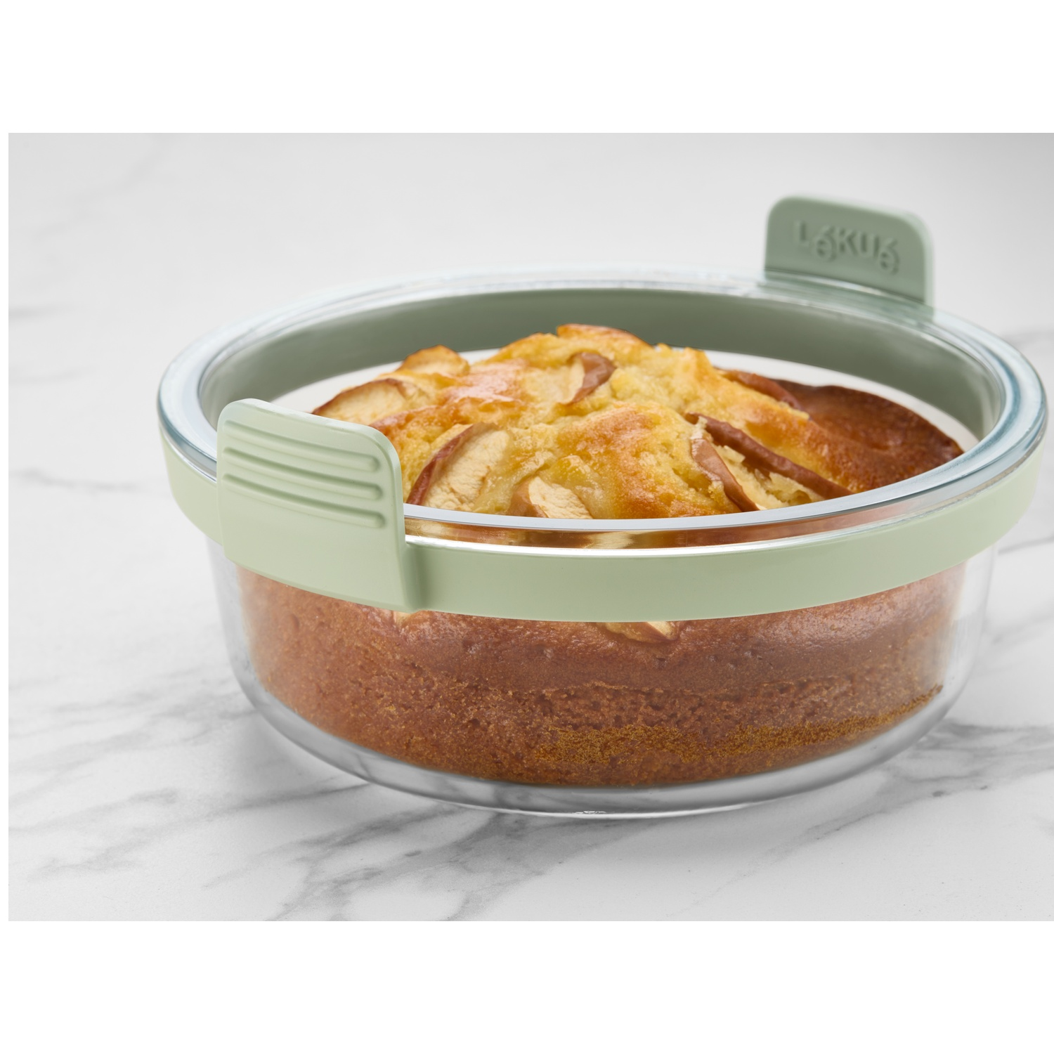 Cake in a glass container with a green lid. The cake has apples on top. It sits on a white and gray marble surface.