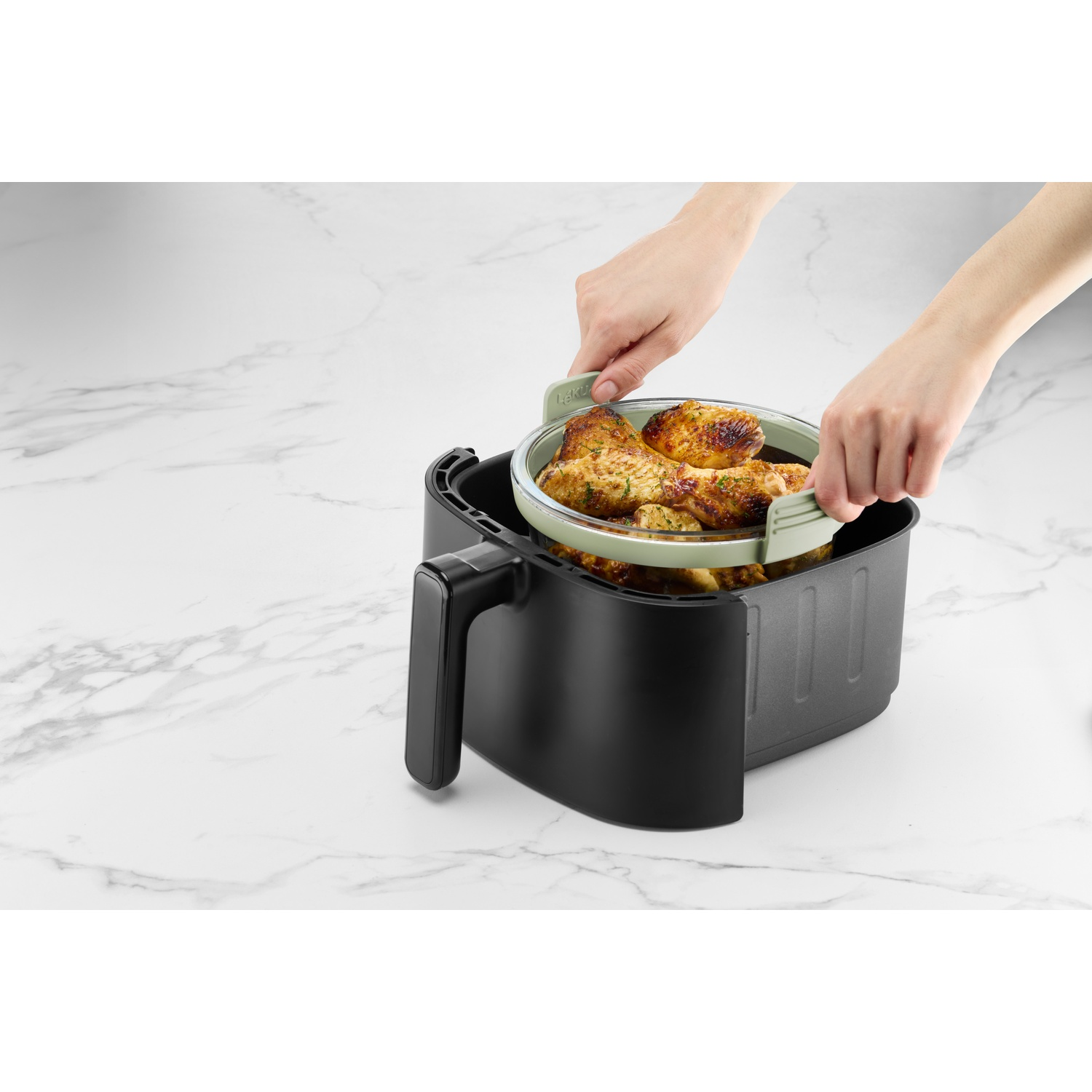 Hands holding a green-rimmed basket of cooked chicken drumsticks, being lowered into a black air fryer on a marble surface.