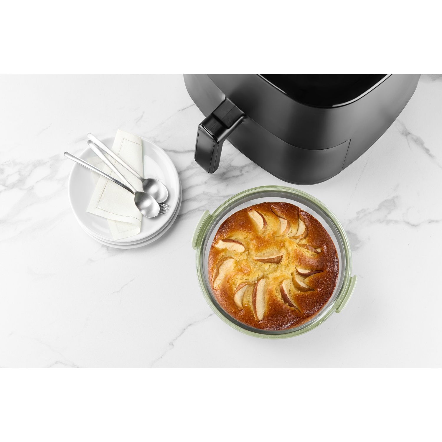 Overhead shot: apple cake in glass container, air fryer, plates with utensils on a marble surface. 