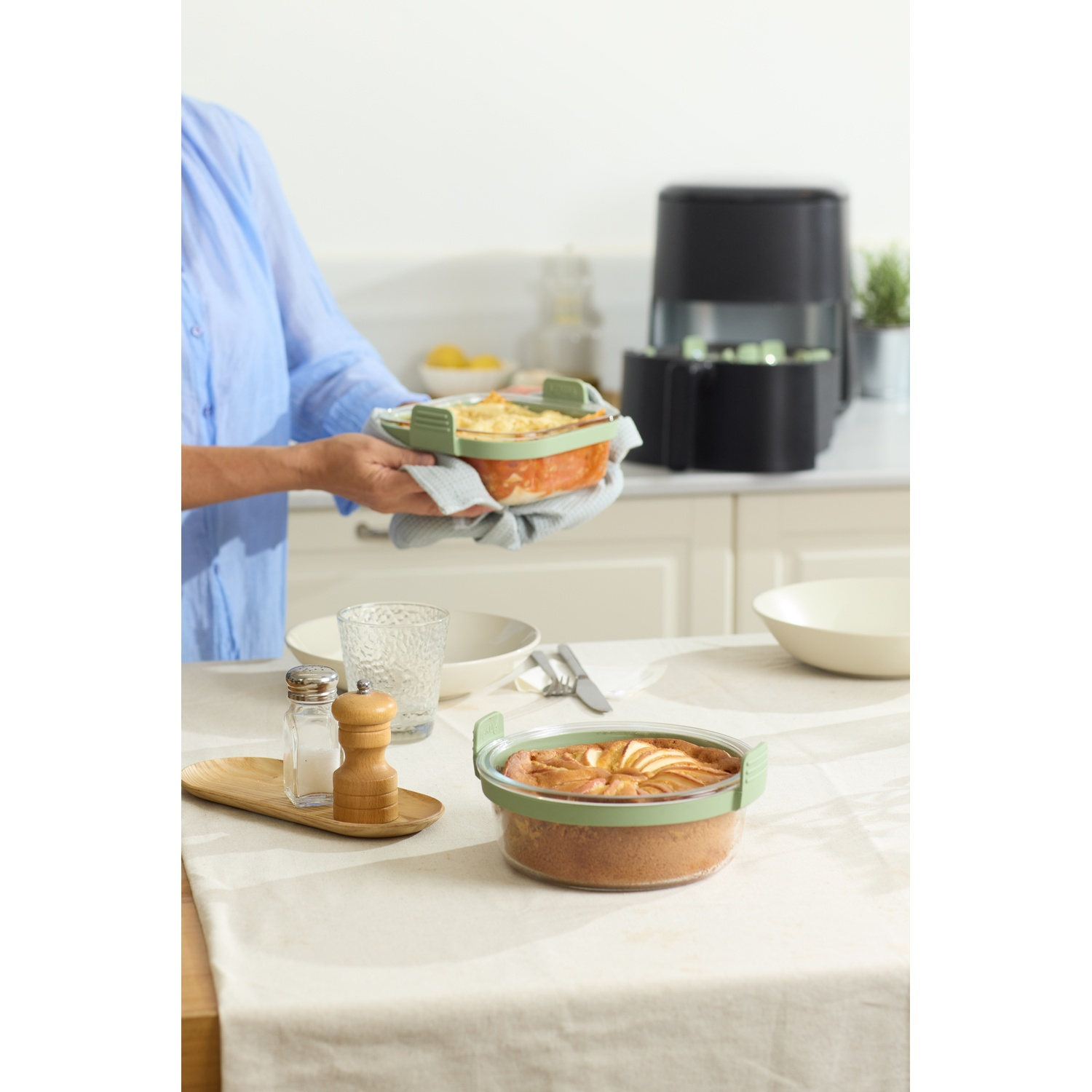 A person holds a casserole dish. A cake is on the table, along with salt and pepper shakers. In the background is a black appliance.