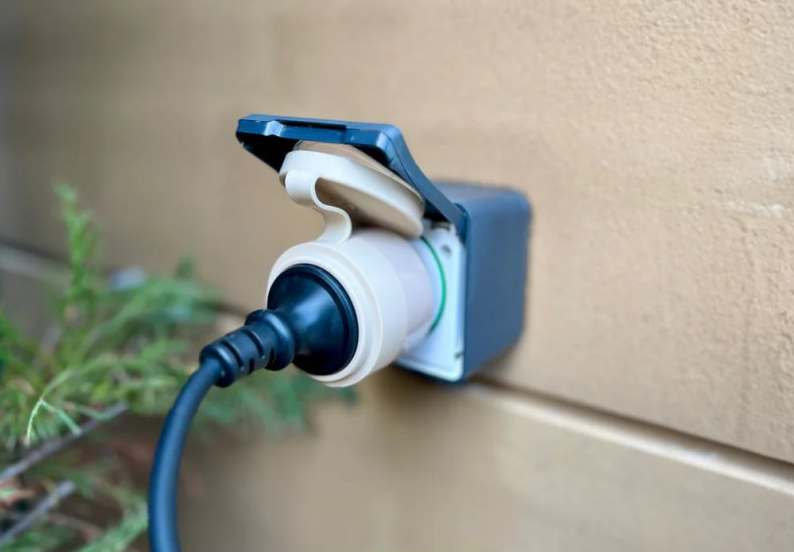 Zewnętrzne gniazdo z czarnym kablem. Gniazdo jest beżowe z niebieską pokrywą, zamontowane na beżowej ścianie. Zielona roślina na pierwszym planie.