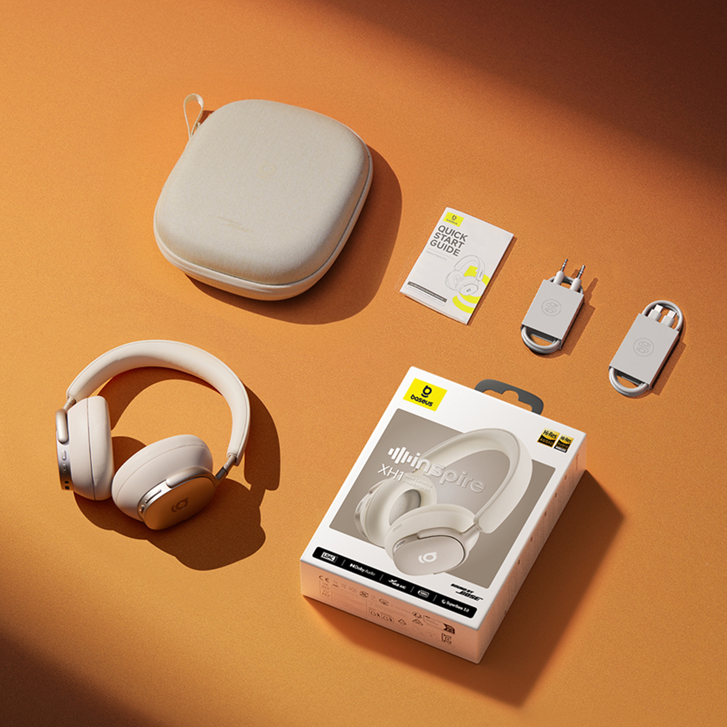 Auriculares de diadema, estuche, guía de inicio rápido y cables de carga sobre una superficie naranja. Los auriculares son blancos y plateados.