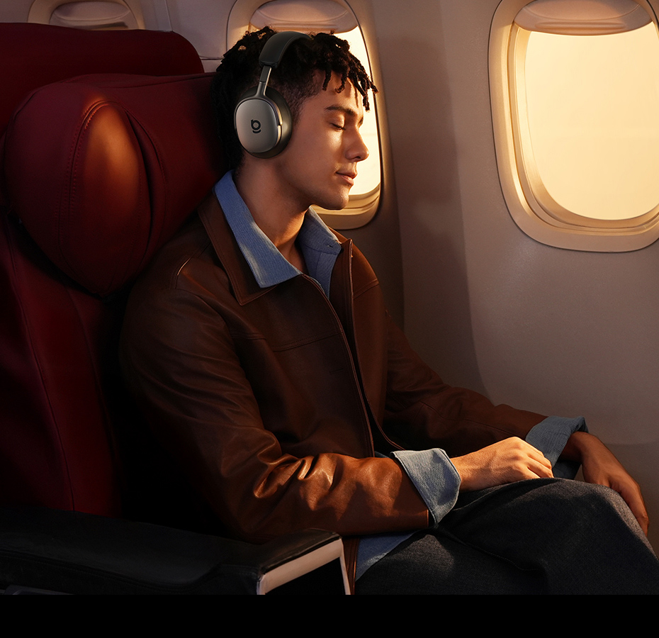 Un hombre con auriculares está sentado en un avión. Lleva una chaqueta marrón y una camisa azul. El asiento es rojo.