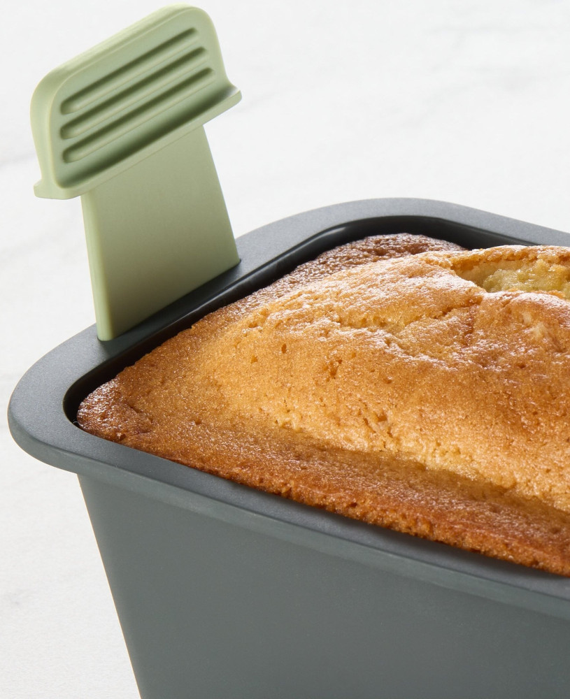 A loaf of cake in a grey baking pan. A green spatula is inserted into the cake. White background.