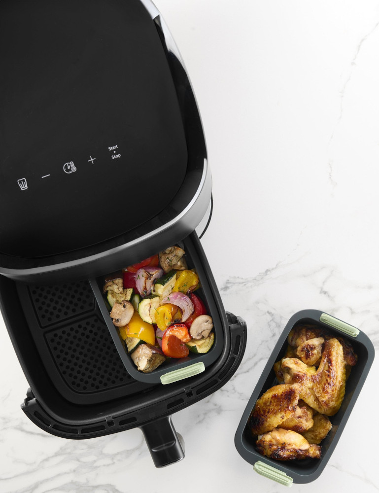 An air fryer with trays of roasted vegetables and chicken wings on a white marble surface.