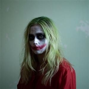 Eine Person mit grünen Haaren und Joker-Make-up trägt ein rotes Hemd. Blasses Gesicht mit dunklen Augen und rotem Lippenstift.