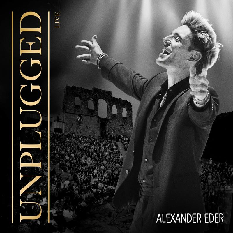 Alexander Eder auf der Bühne, Arme erhoben. Publikum im Hintergrund. Text Unplugged Live links.