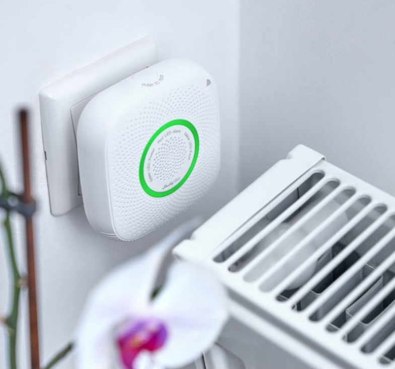 Biały czujnik dymu jest podłączony do gniazdka ściennego. Obok biały grzejnik. Na pierwszym planie biała orchidea.