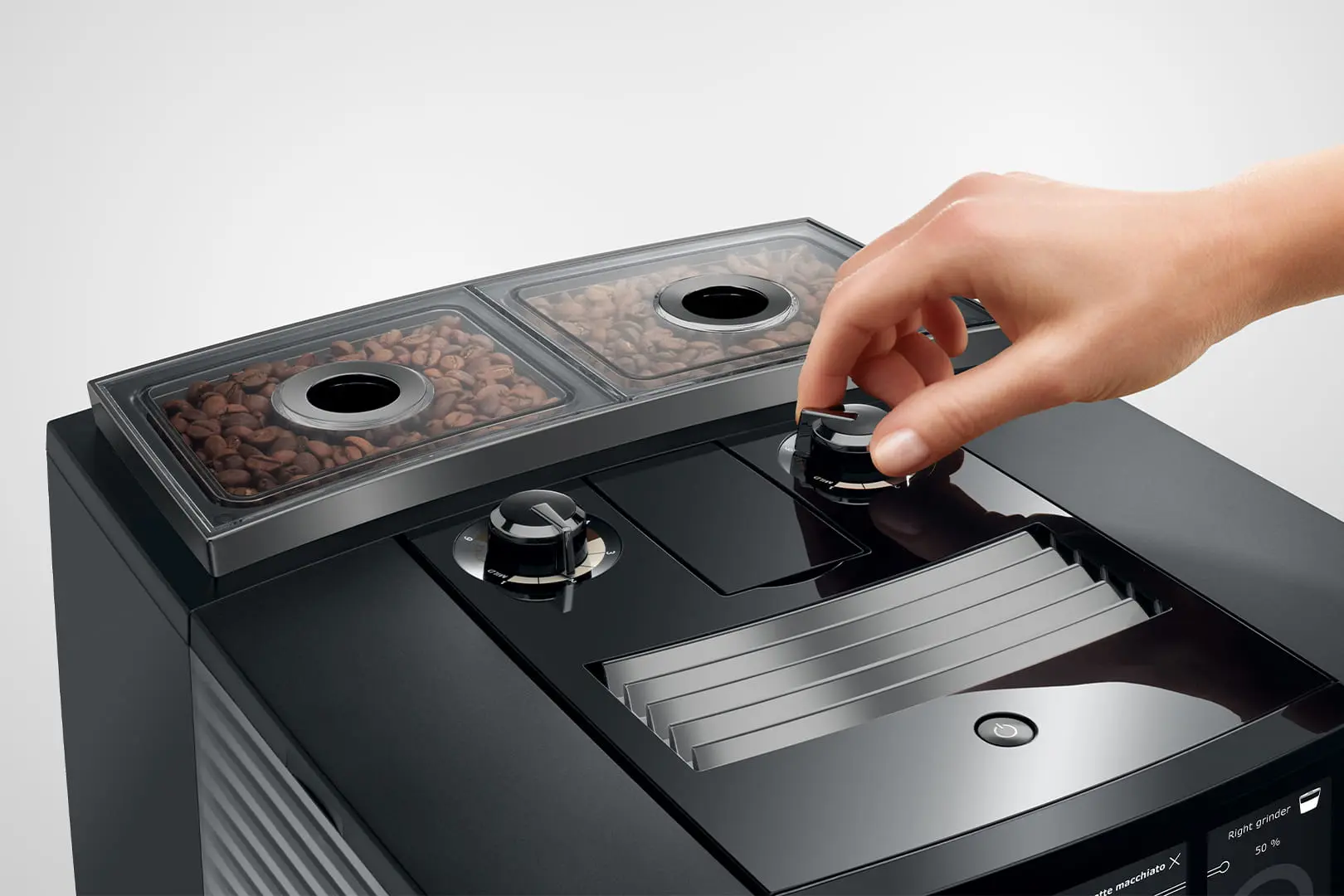 Eine Hand dreht einen Drehregler an einer schwarzen Kaffeemaschine. Bohnen sind in einem durchsichtigen Behälter sichtbar. Der Hintergrund ist weiß.
