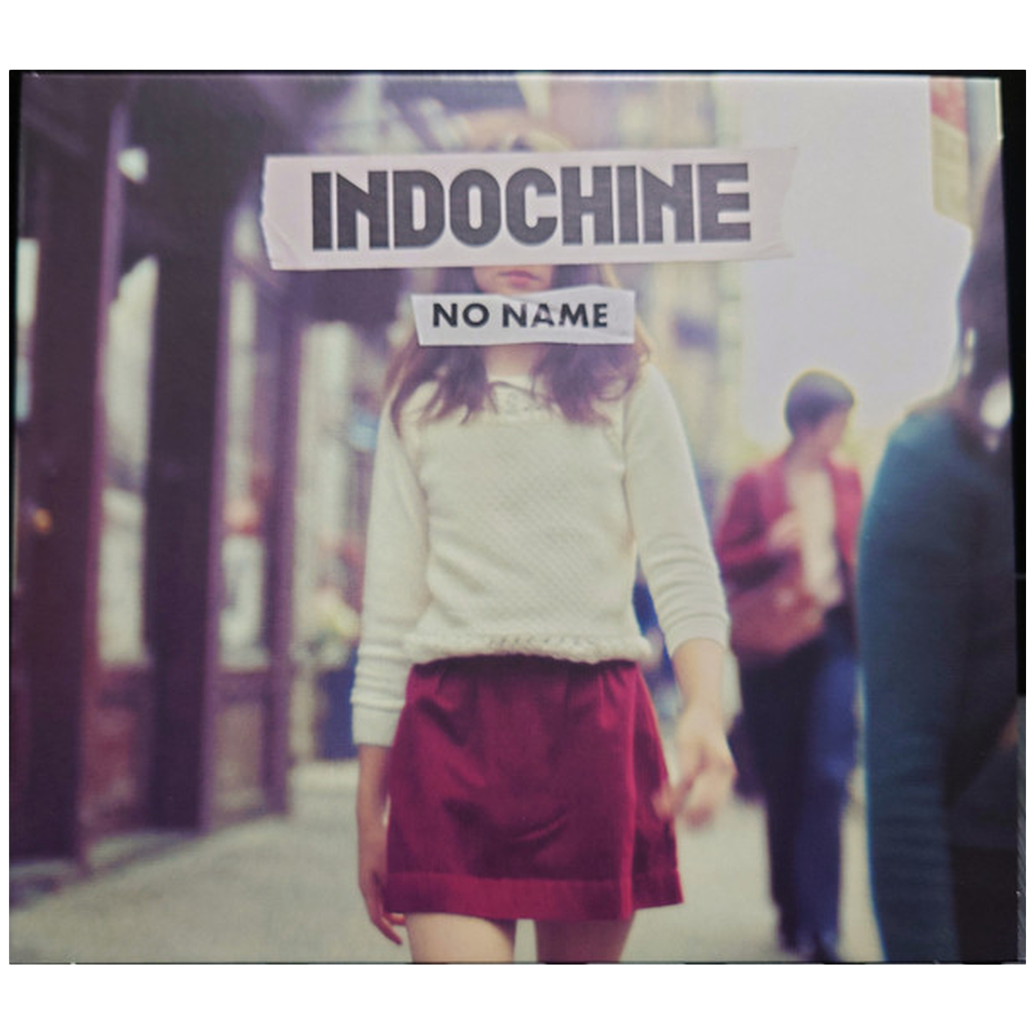 Une personne en pull blanc et jupe rouge marche dans la rue. Le mot 'Indochine' est au-dessus de ses yeux et 'No Name' en dessous.