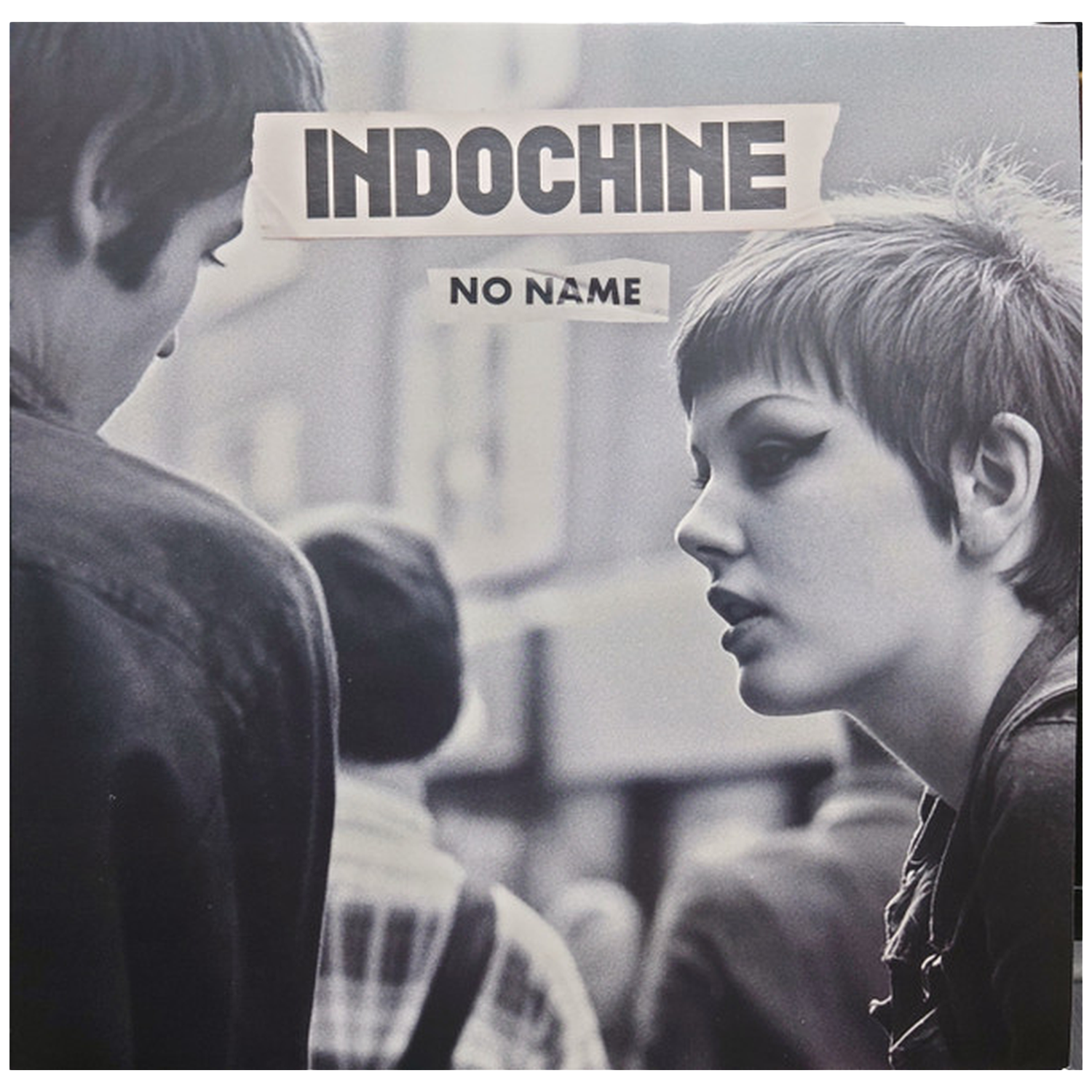 Zwart-wit foto. Een vrouw met kort haar en donkere eyeliner kijkt naar rechts. De rug van een man is links. Tekst: INDOCHINE, NO NAME.