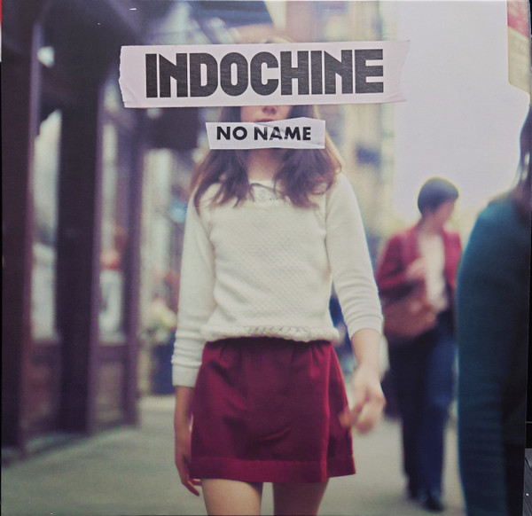 Une personne marche dans une rue. Le visage est couvert par le texte 'INDOCHINE NO NAME'. Elle porte un pull blanc et une jupe rouge.