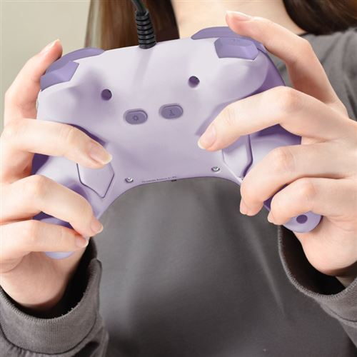 Una persona sostiene un mando de juegos morado con ambas manos. El mando tiene botones y un cable. La persona lleva una camisa gris.