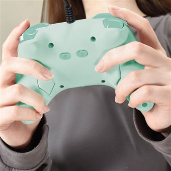 Manos sosteniendo un mando de juego verde claro con botones y un cable negro. Camisa gris al fondo.