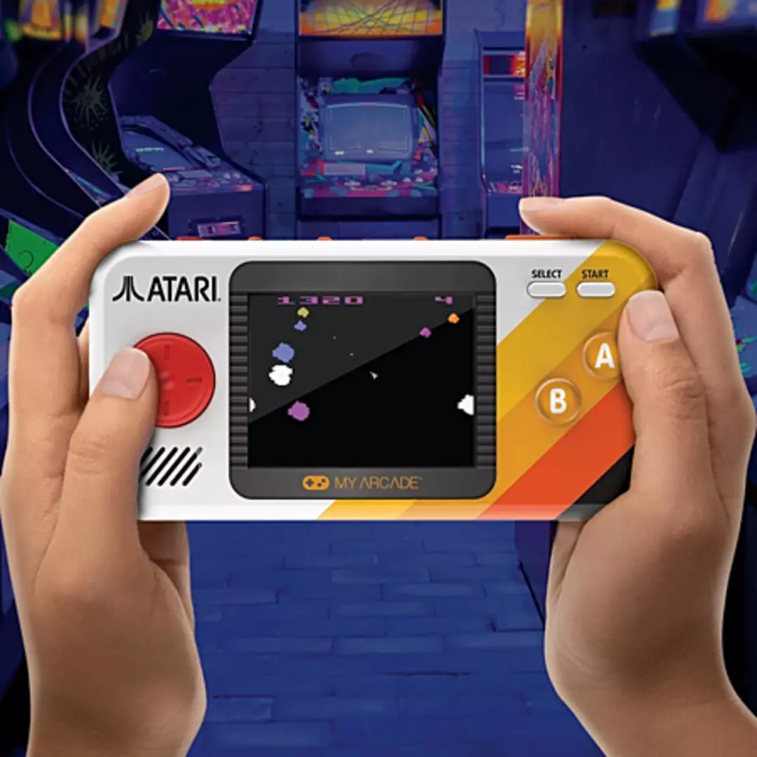 Manos sosteniendo una consola de juegos portátil retro. La pantalla muestra un juego, con máquinas recreativas al fondo.