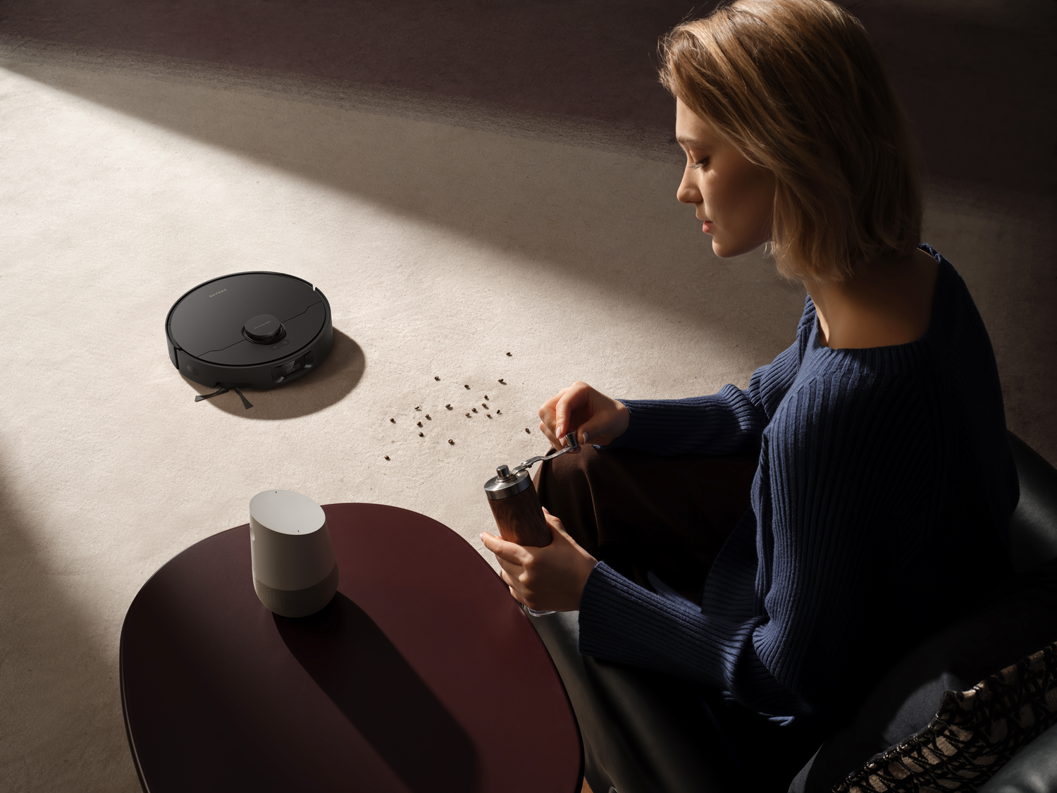 Una mujer muele especias junto a un altavoz inteligente y un robot aspirador. La escena está iluminada por luz natural.