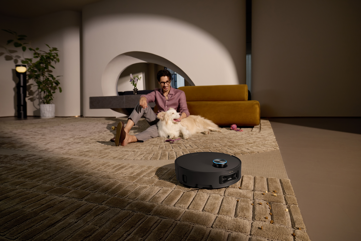 Un hombre acaricia a un perro en una alfombra, cerca hay un robot aspirador. En el fondo hay un sofá amarillo.