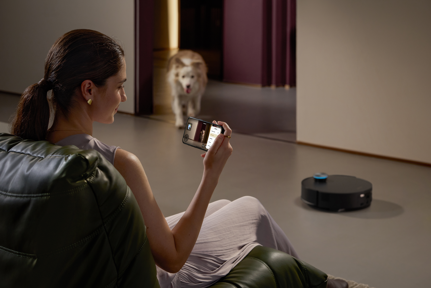 Una mujer usa su teléfono, mirando a un perro en la puerta. Una aspiradora robot limpia el suelo. Silla verde en primer plano.
