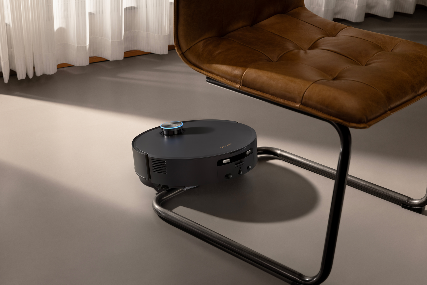 Una aspiradora robot negra se encuentra debajo de una silla de cuero marrón, sobre un suelo gris claro. Una cortina blanca transparente está en el fondo.