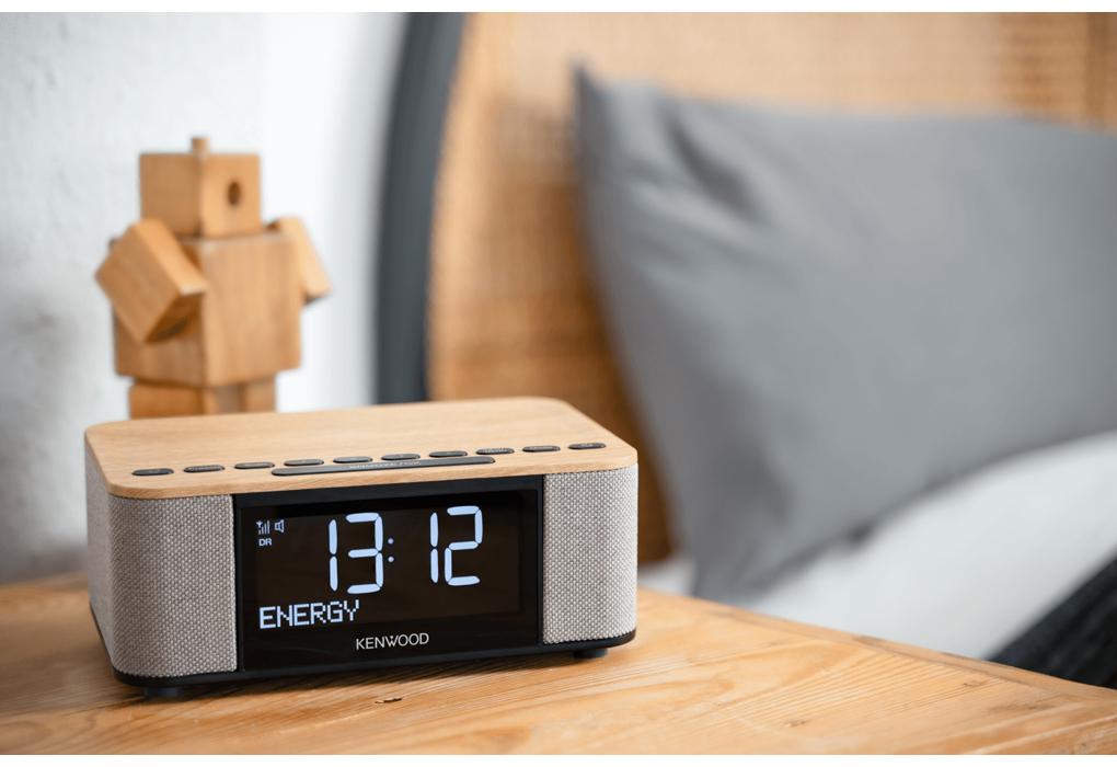 Eine Holzuhr mit Anzeige 13:12 auf einem Holztisch. Ein Holzroboter und ein graues Kissen im Hintergrund.
