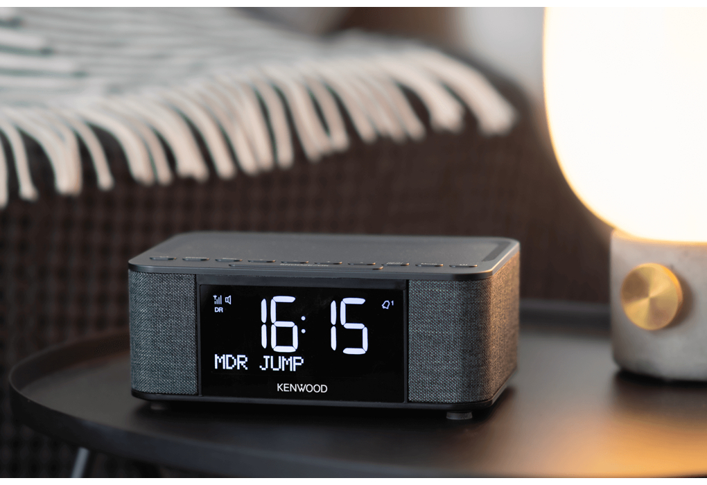 Eine Digitaluhr mit der Anzeige 16:15 steht auf einem schwarzen Tisch. Eine Lampe und ein verschwommener Hintergrund sind sichtbar.