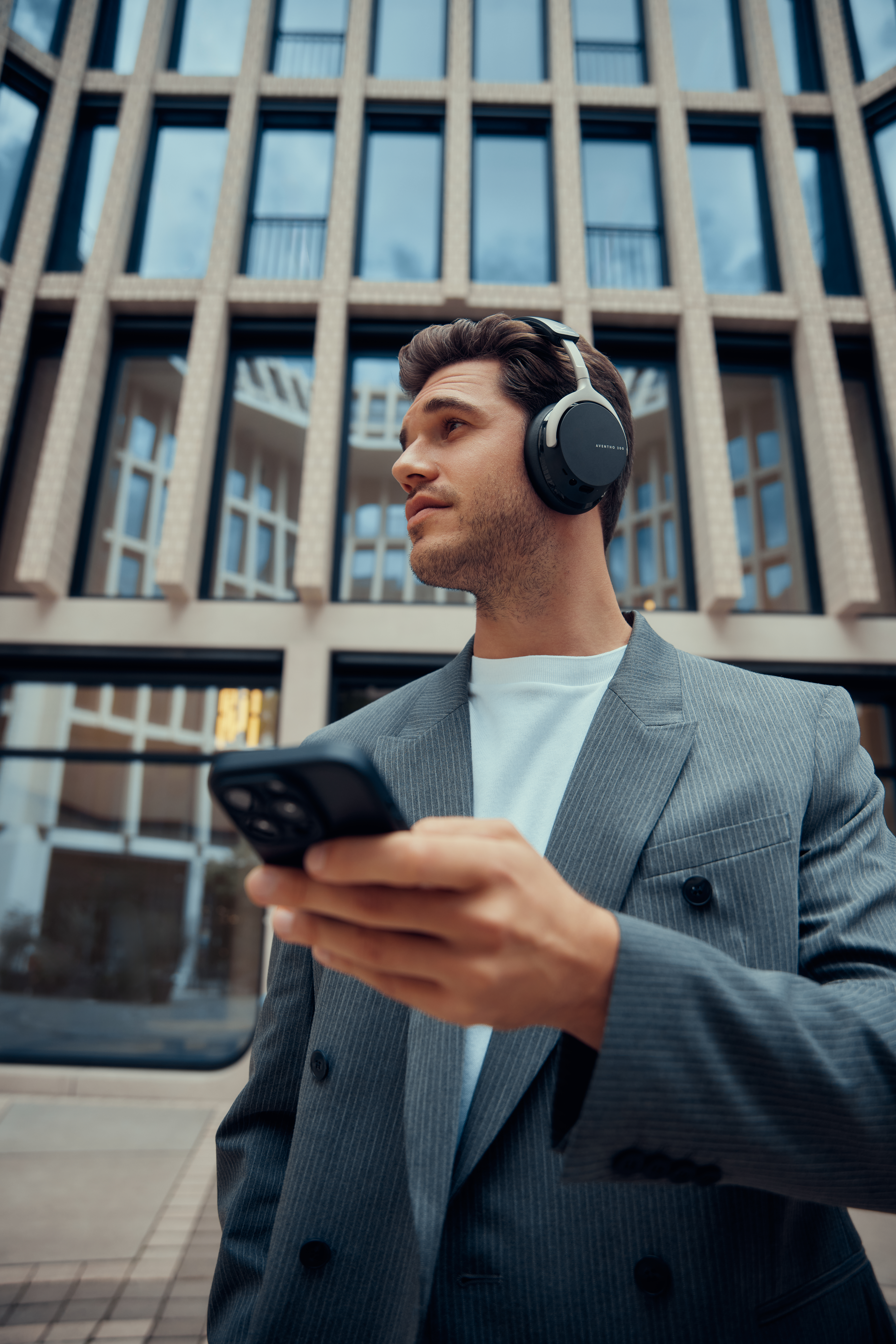 Ein Mann im Anzug mit Kopfhörern und Telefon, der nach oben schaut, vor einem modernen Gebäude stehend.
