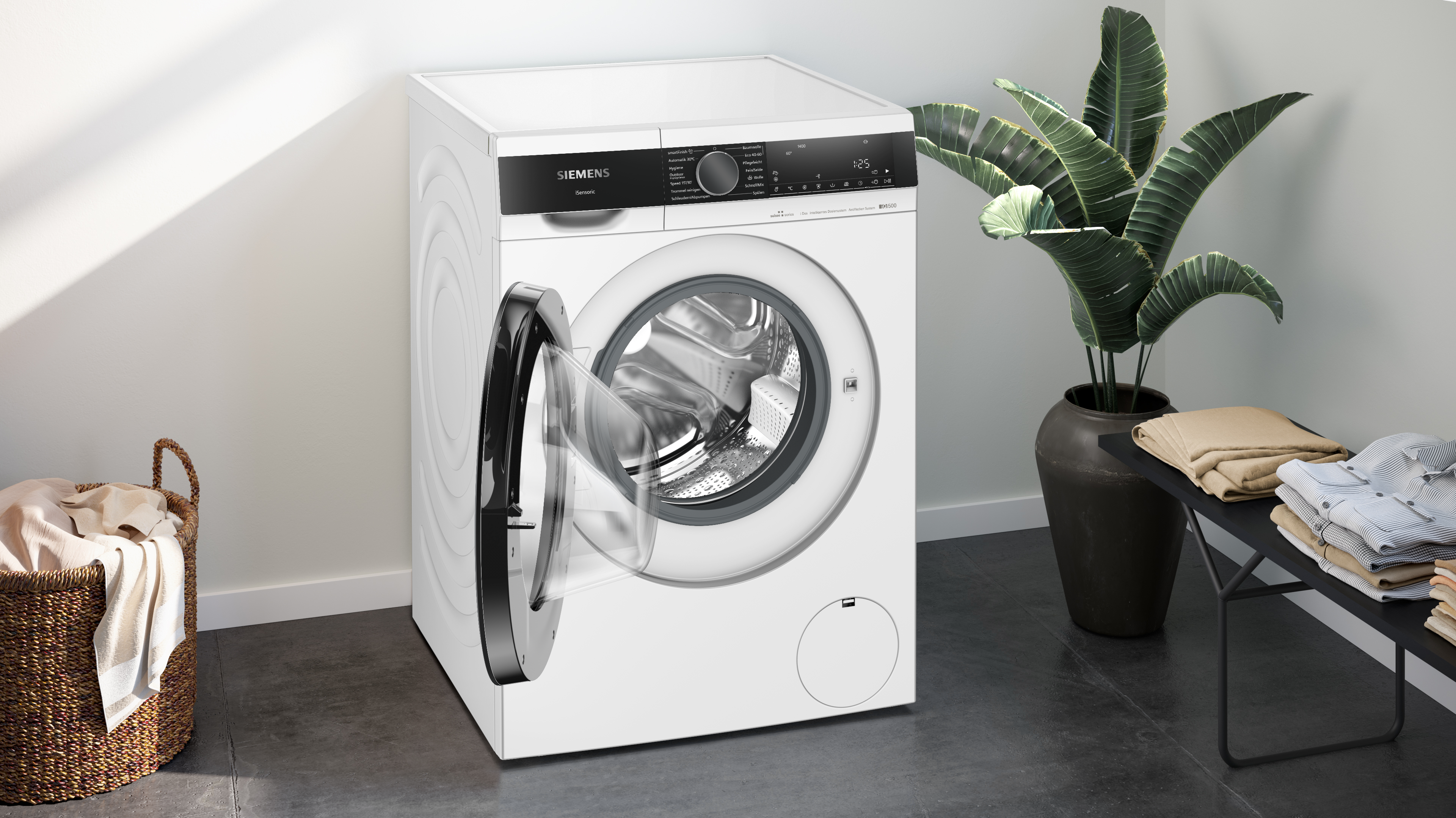 Una lavatrice Siemens bianca con porta aperta. Un cesto di vestiti e una pianta ai lati.