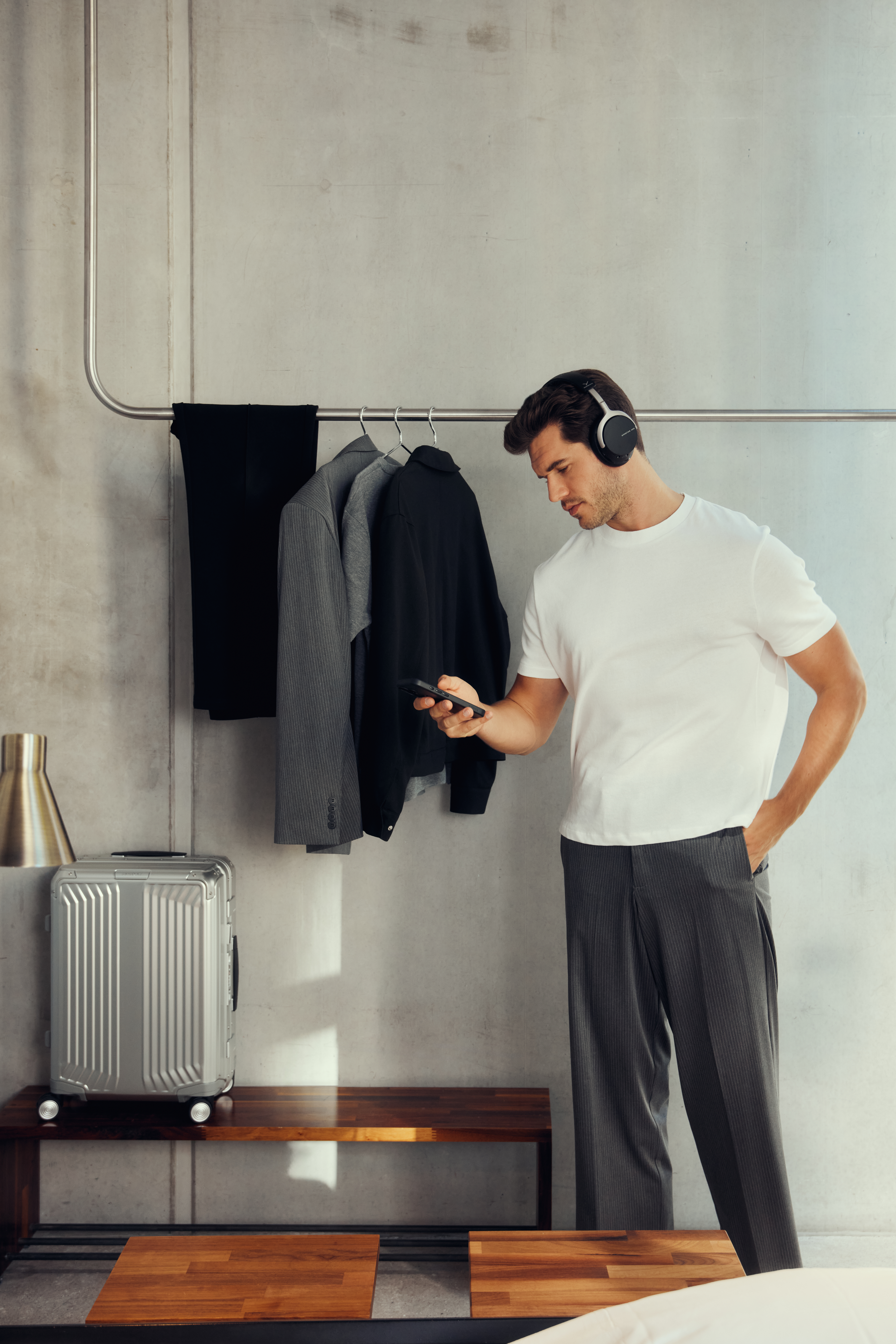 Mann mit Kopfhörern, blickt auf das Telefon. Kleidung hängt hinter ihm. Silberner Koffer, Lampe und Bank sind sichtbar.