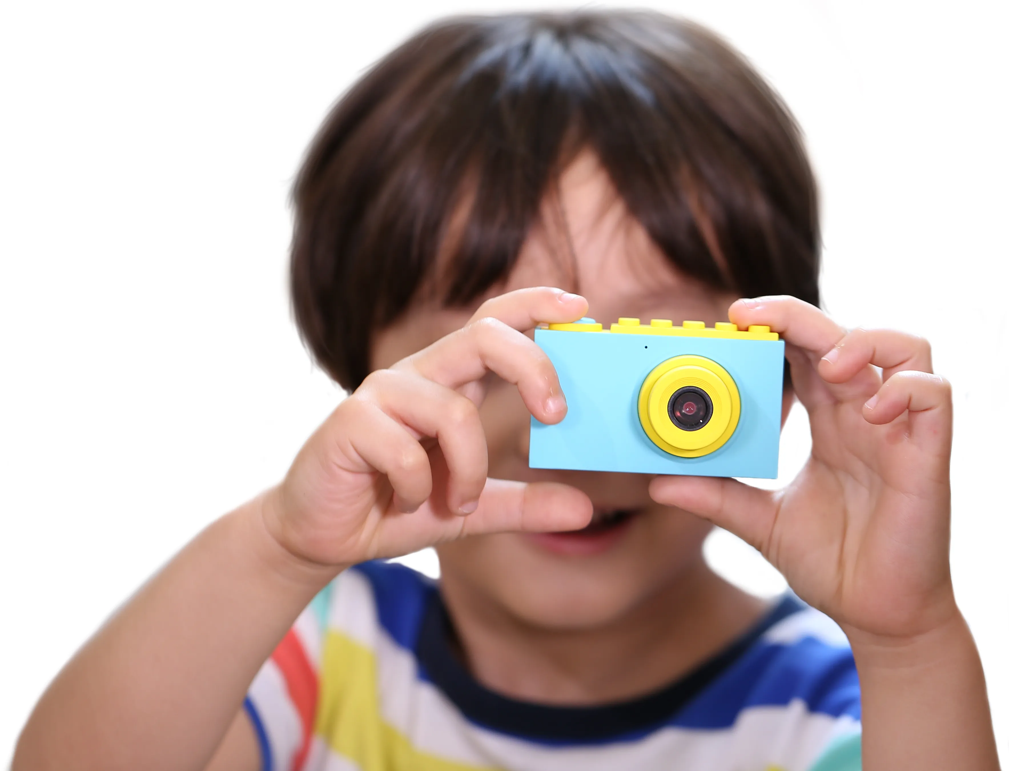 Un enfant tient un appareil photo jouet bleu et jaune. Le visage de l'enfant est partiellement visible. Fond blanc.