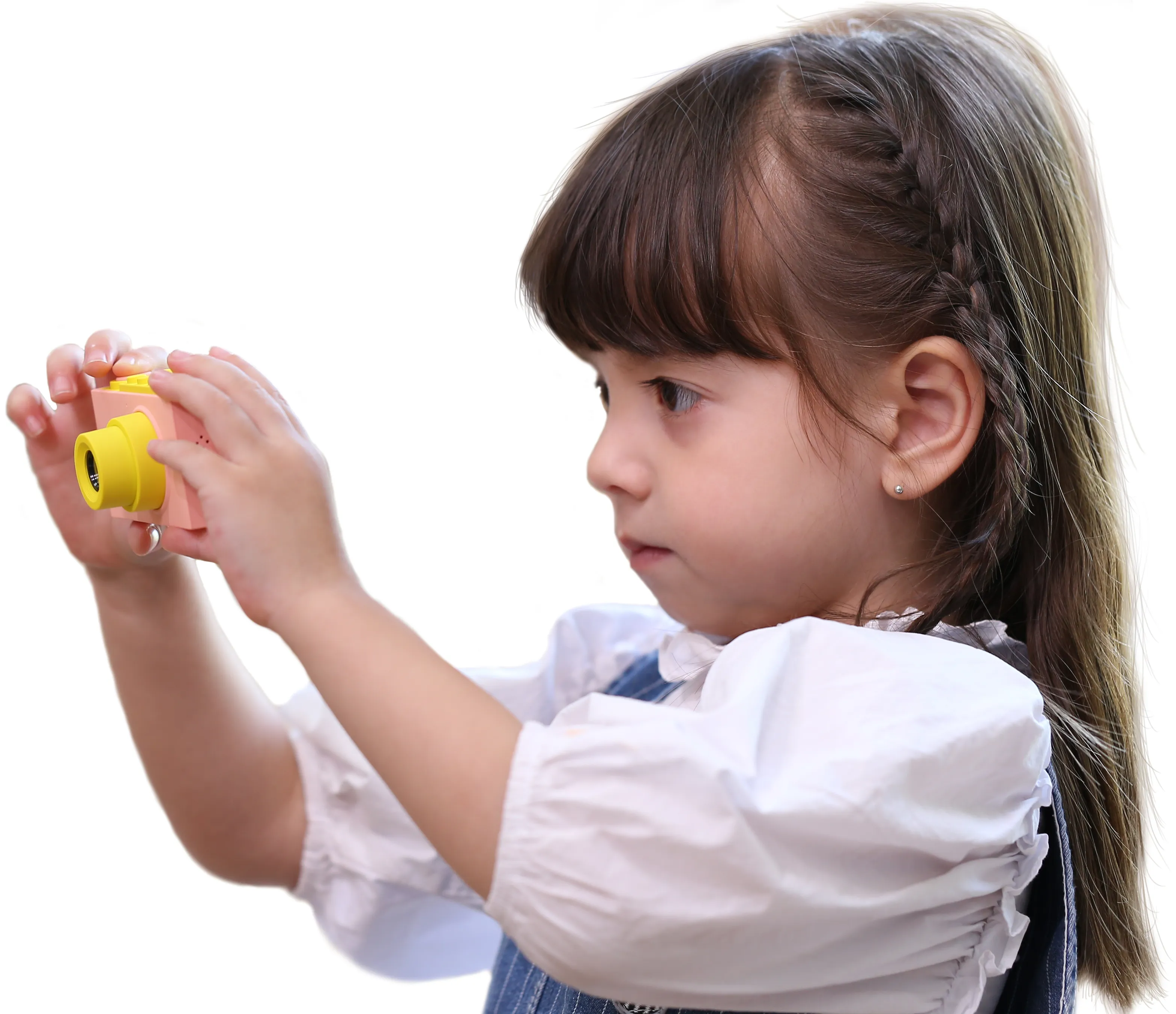 Une jeune fille tient un appareil photo jouet rose et jaune, en regardant à travers. Elle porte une chemise blanche et une salopette en jean.