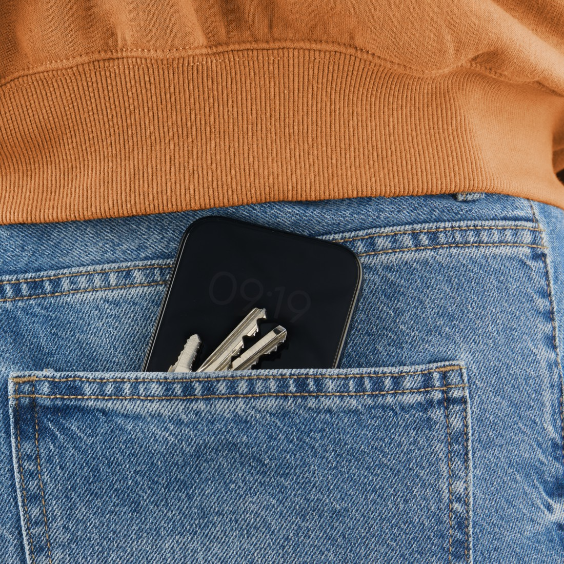 Nahaufnahme des Rückens einer Person, die eine blaue Jeans-Tasche mit Handy und Schlüsseln zeigt. Das Telefon zeigt die Uhrzeit 09:19 an.