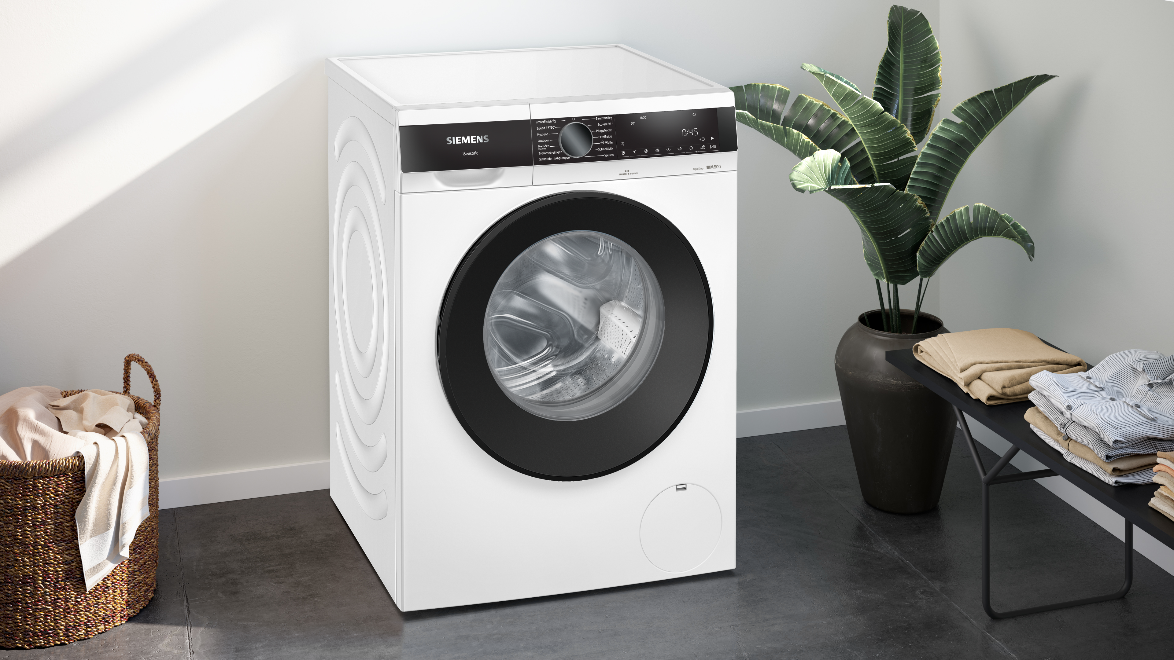 Eine weiße Siemens-Waschmaschine mit schwarzer Tür. Rechts eine Pflanze in einem dunklen Topf. Wäschekorb links.