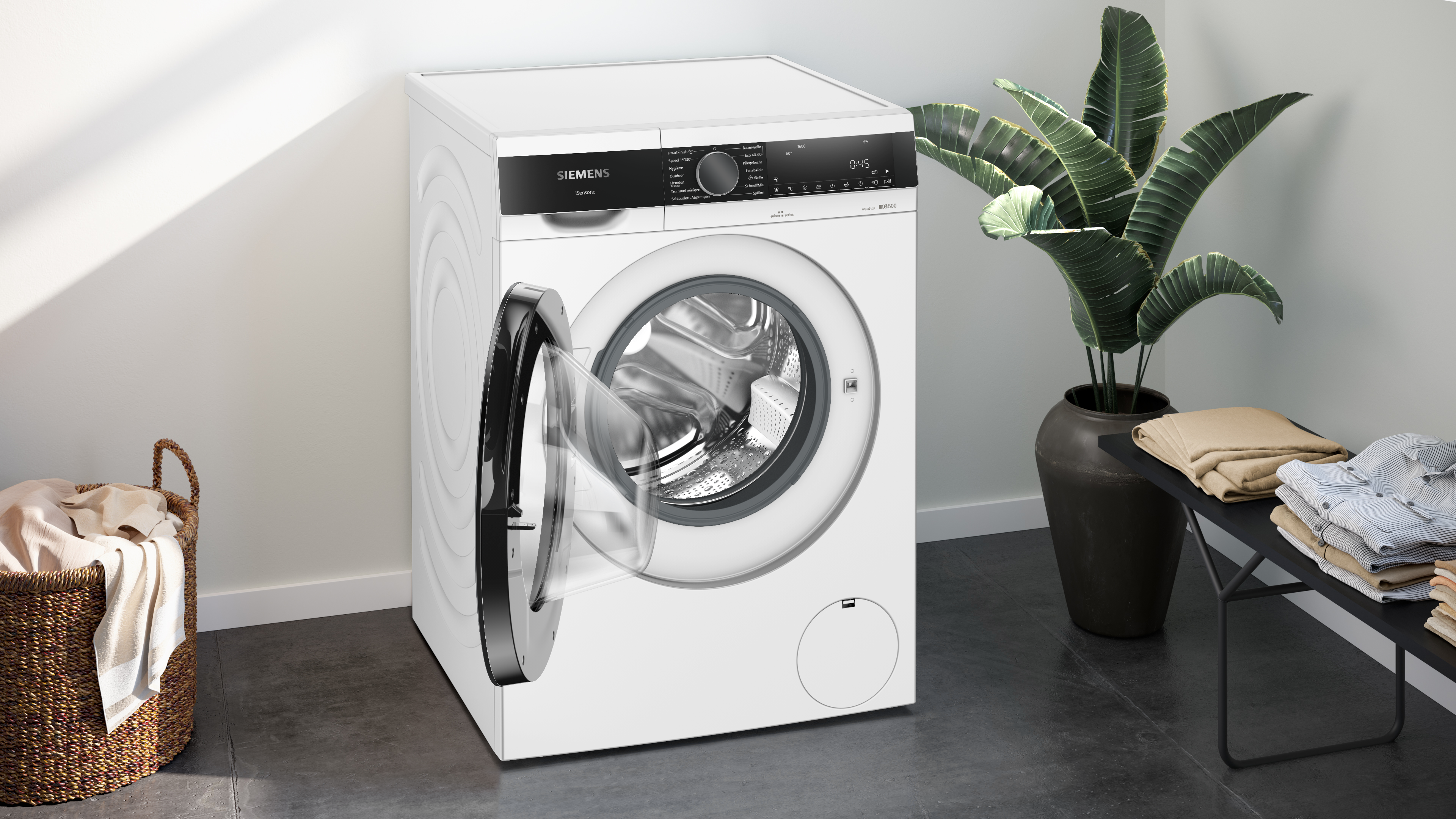 Eine weiße Siemens Waschmaschine mit offener Tür. Wäschekorb links, Pflanze rechts und gefaltete Kleidung.